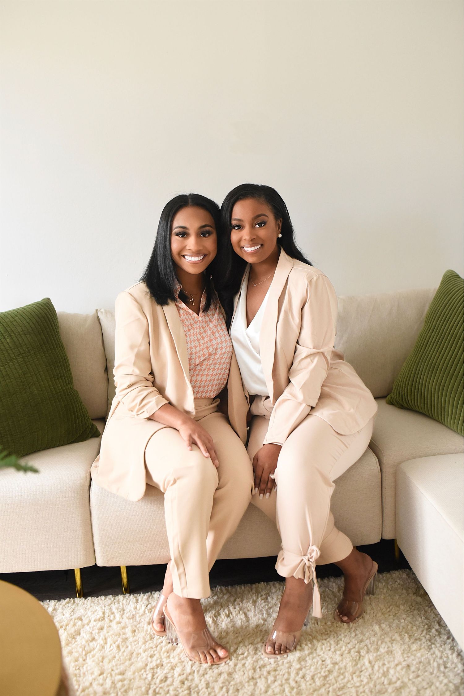 Two women in matching beige suits sitting together on neutral sofa with green pillows.