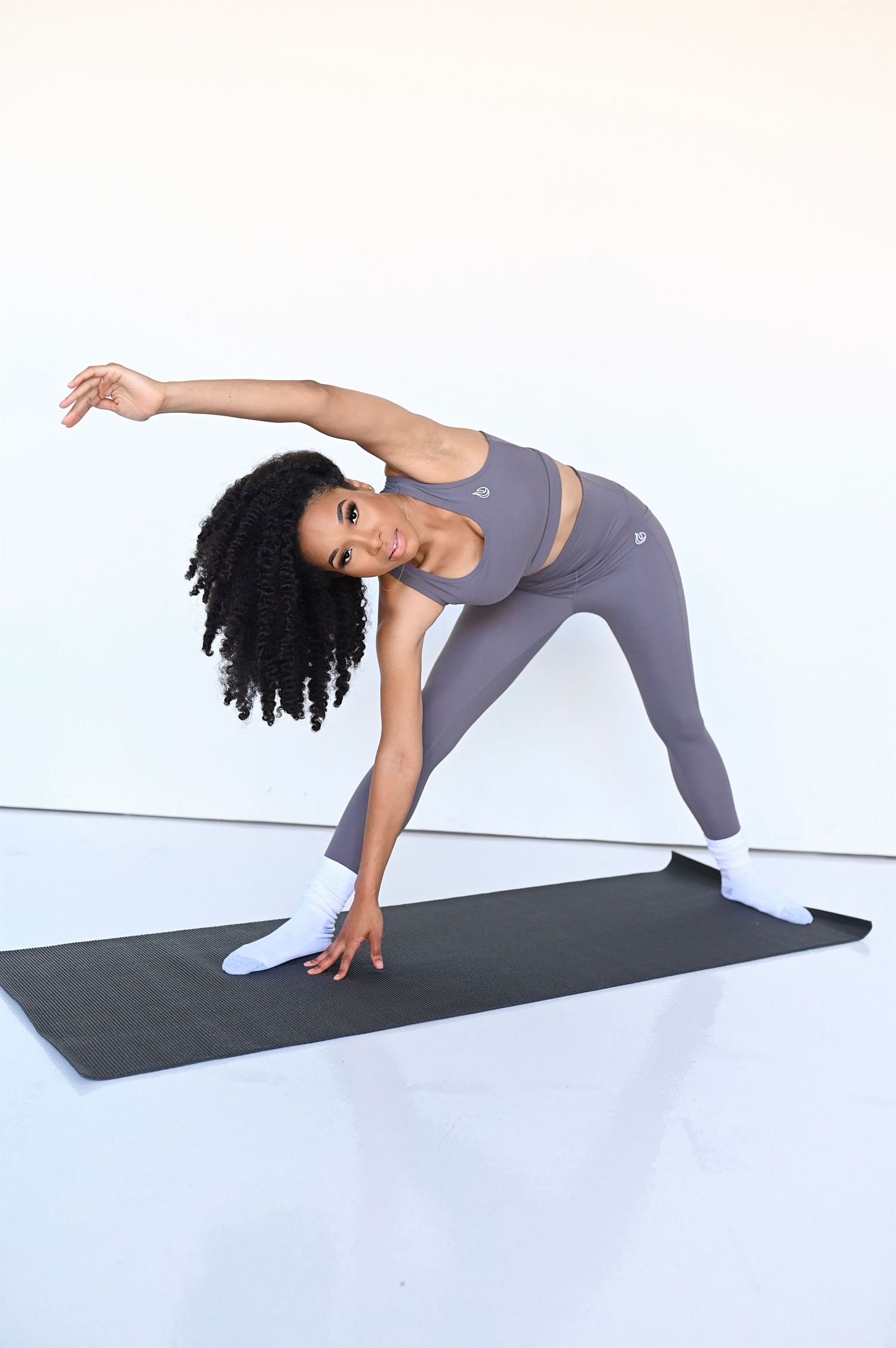 Athletic woman in gray workout set performs a triangle yoga pose on a black mat against white background.