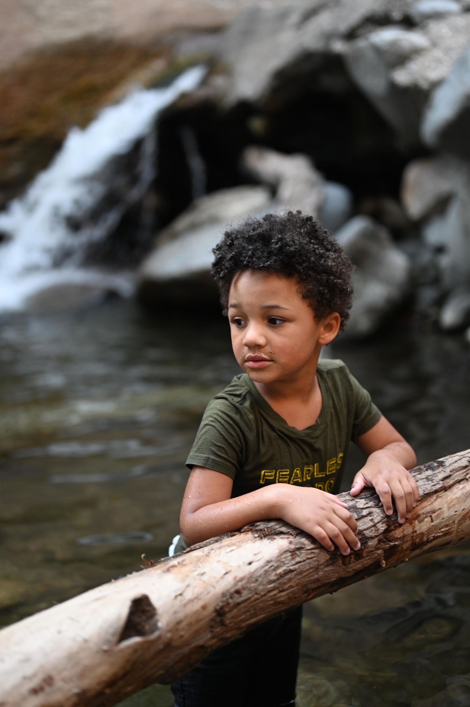 Young Black boy leaning on fallen log near rocky waterfall stream in green shirt.