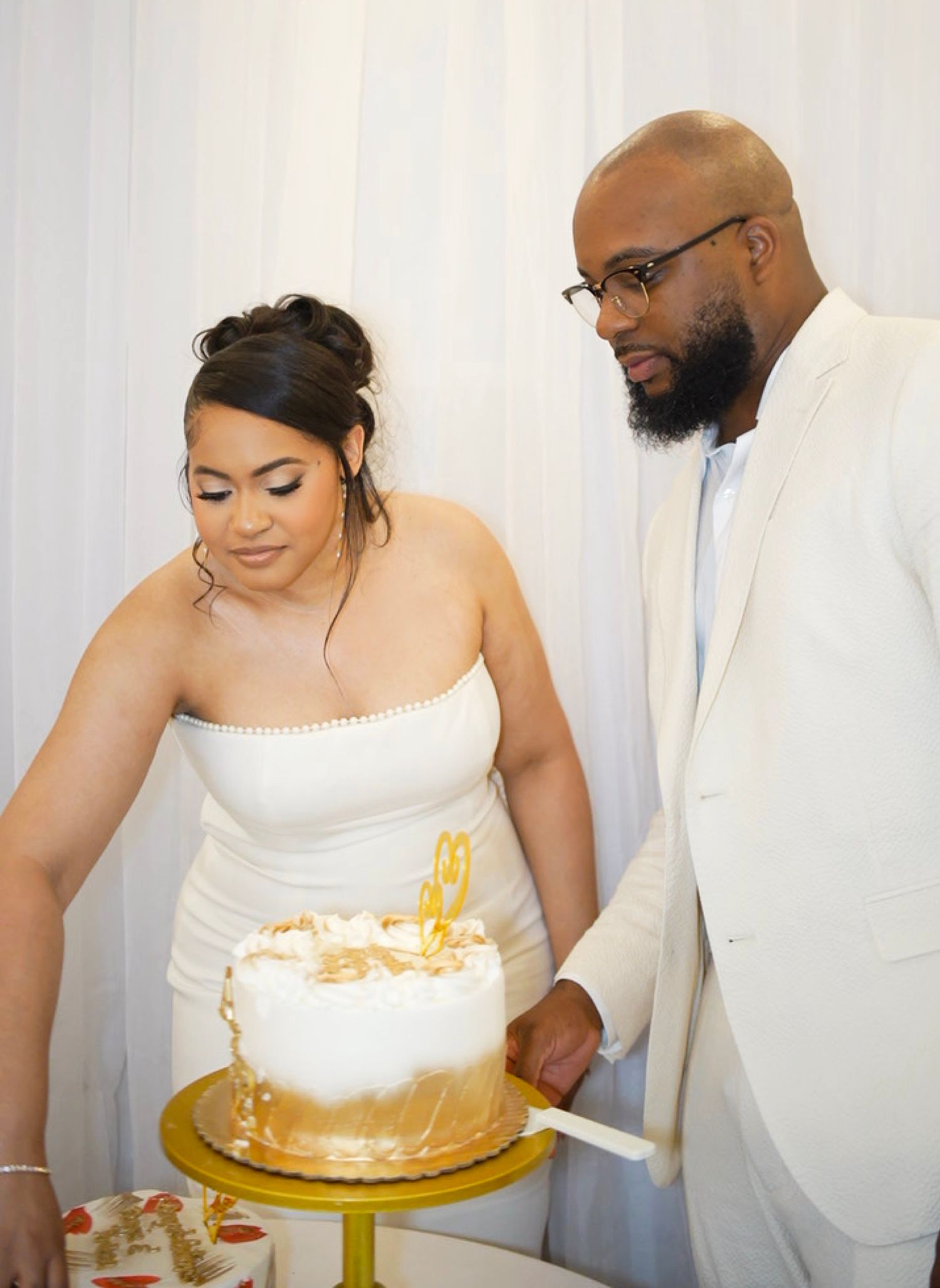 Couple in white attire cutting a gold and white wedding cake together.