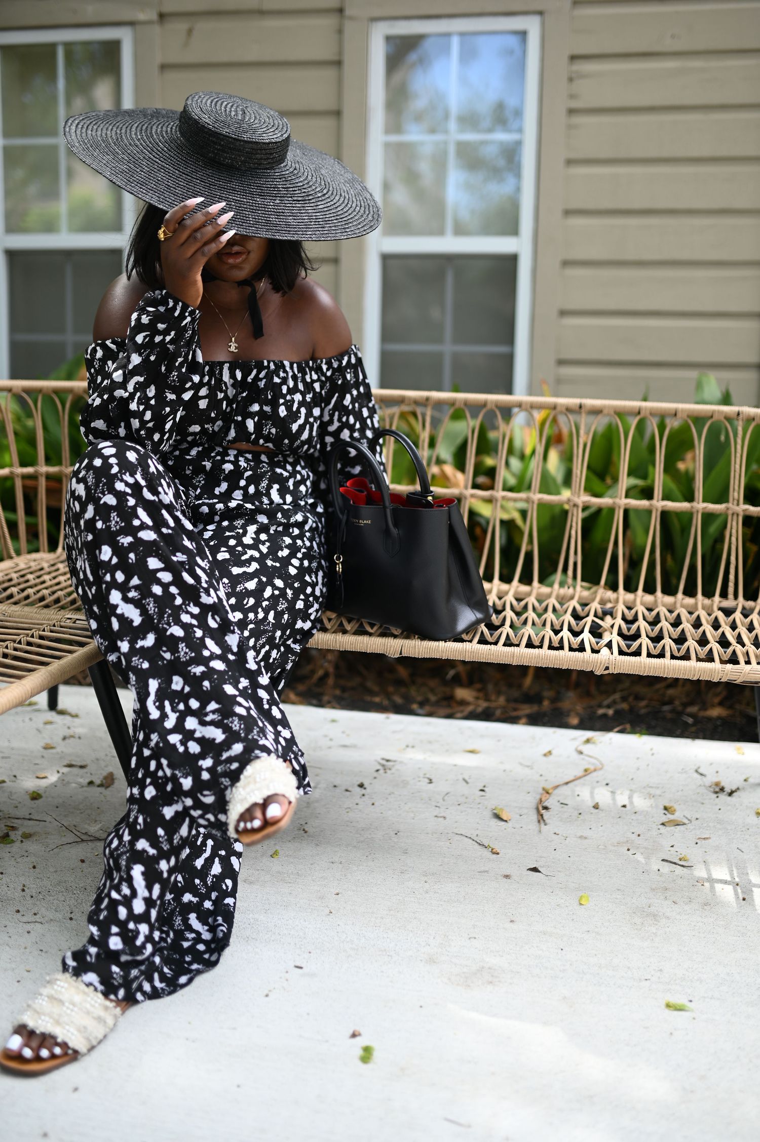 Woman in black and white printed off-shoulder jumpsuit and wide brim hat sits on rattan bench.