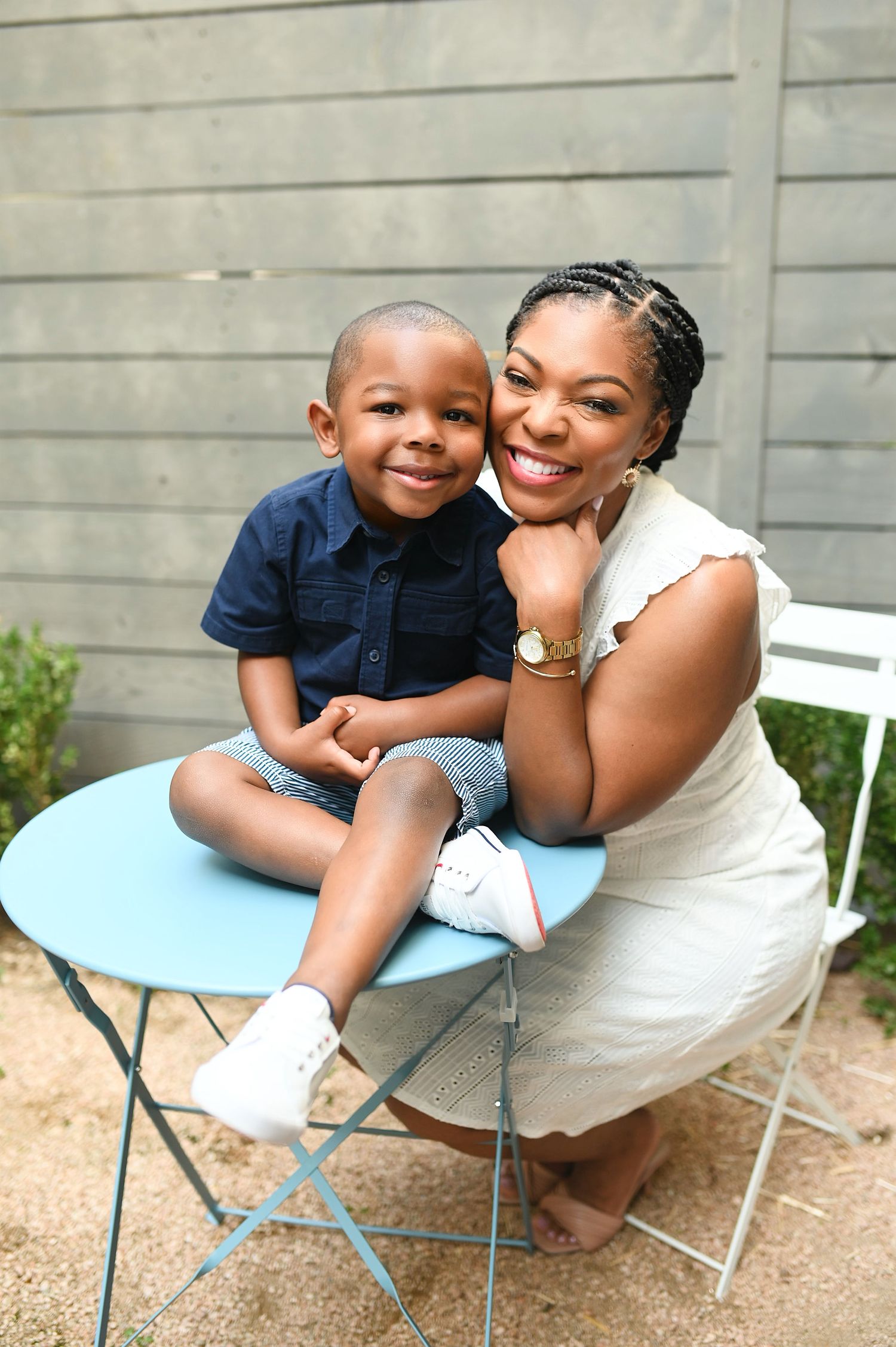 A smiling Black mother and young son pose together at a blue outdoor table.