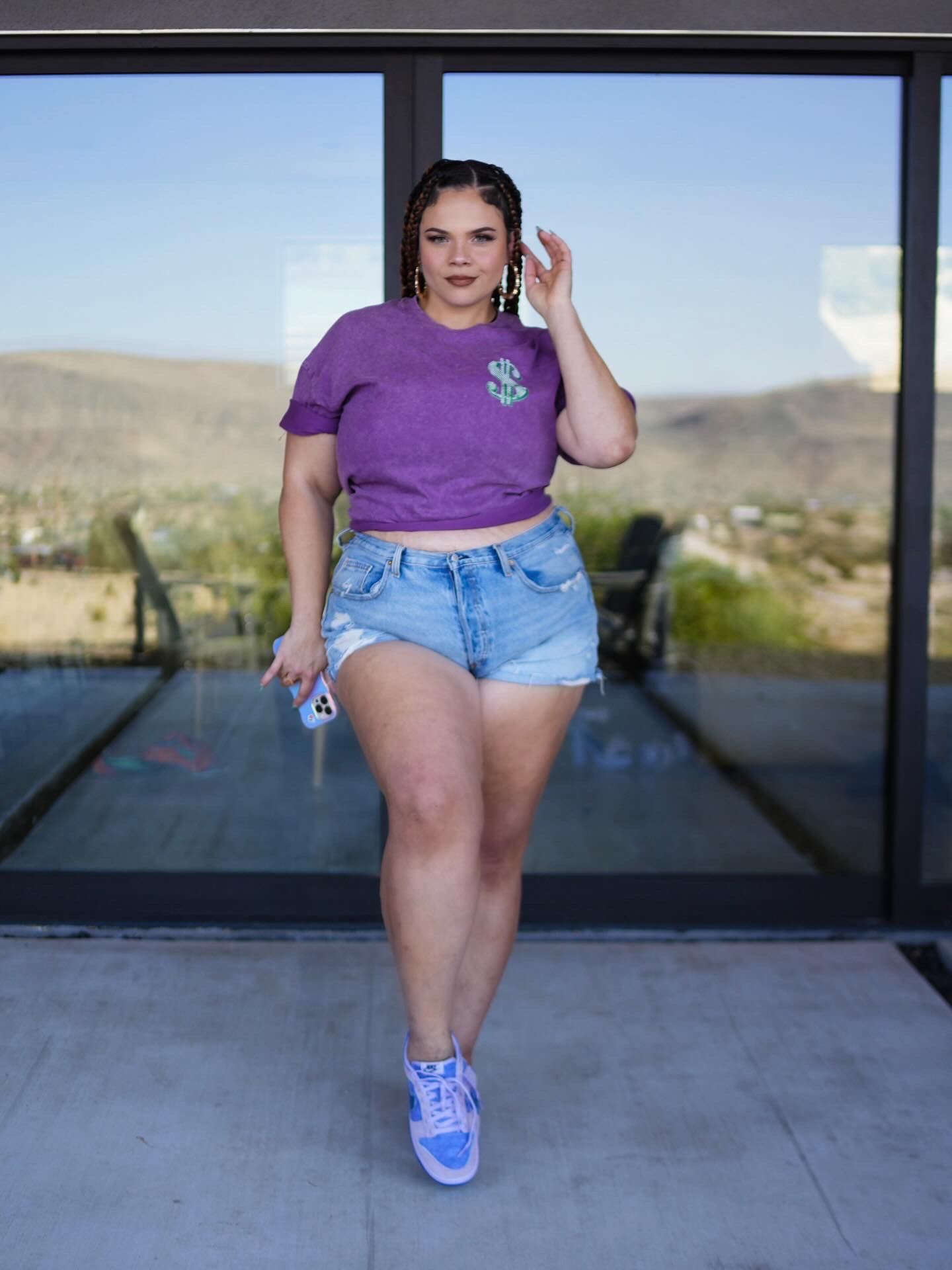 Woman in purple graphic crop tee, denim cutoff shorts, and lavender Air Jordans poses on a desert home patio.