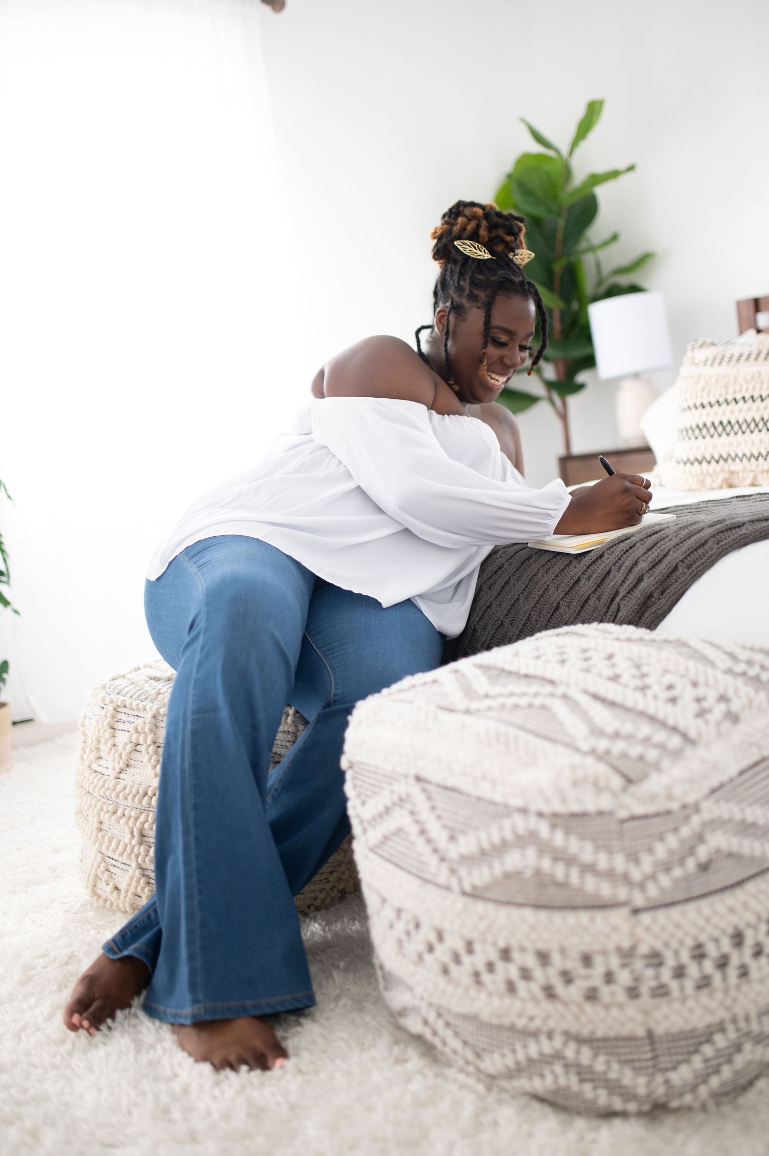 Woman in white off-shoulder top and flared jeans sits on bed writing in notebook.