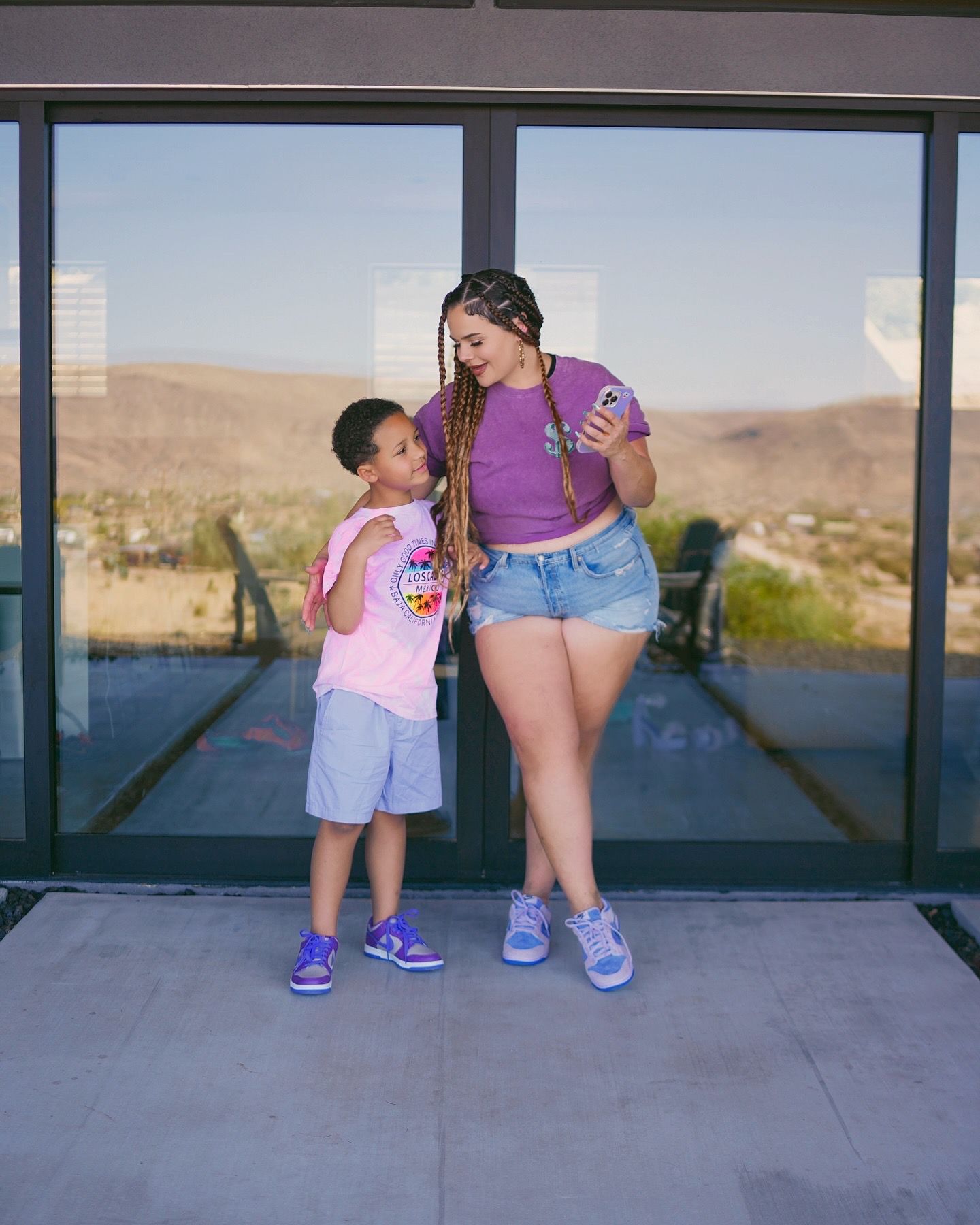 Woman in purple crop top and denim shorts showing phone to young child in pink outfit near glass doors.