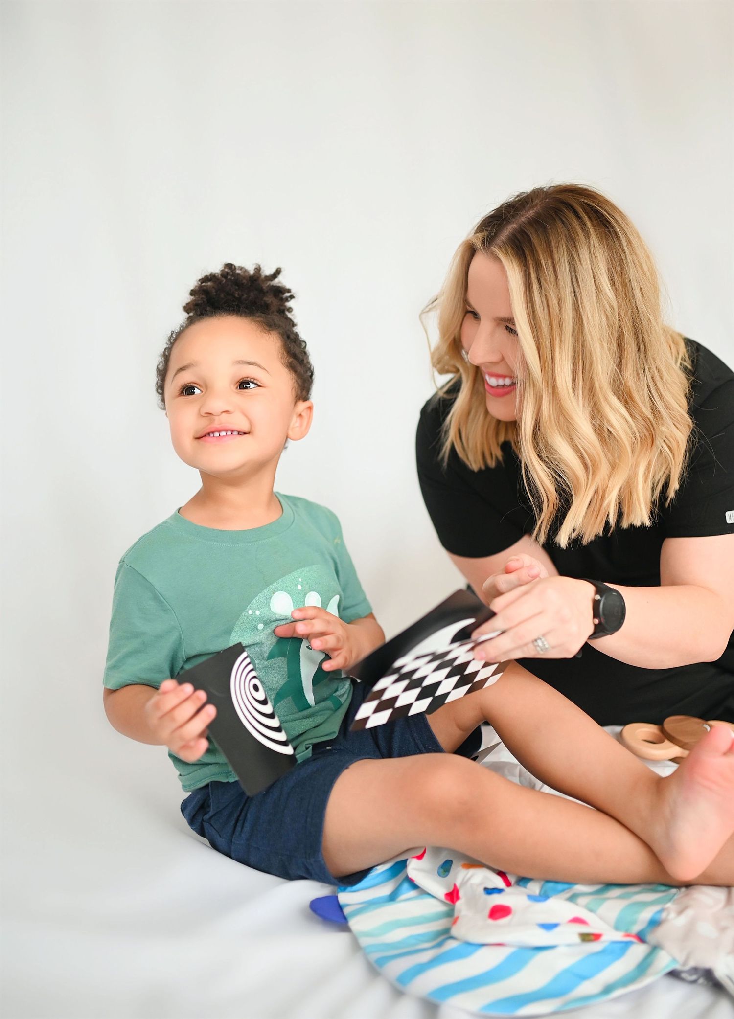 Smiling blonde woman sits with an excited young boy holding black and white sensory cards on a white surface.