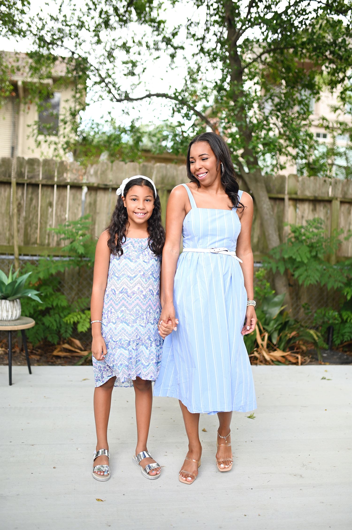 Woman and young girl in coordinating blue and white dresses holding hands in outdoor backyard setting.