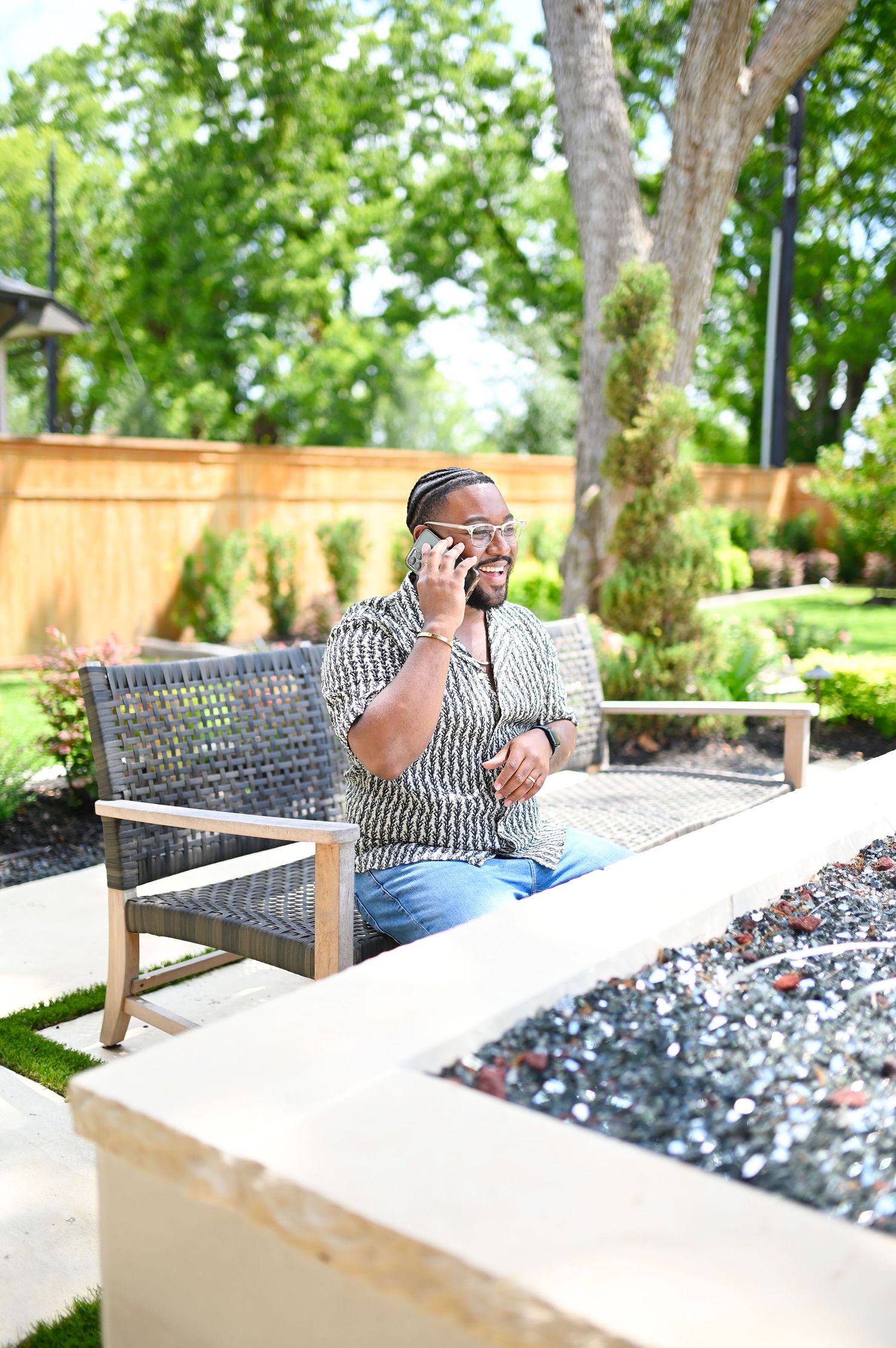 Man in patterned shirt talking on phone while seated on bench in a lush garden.