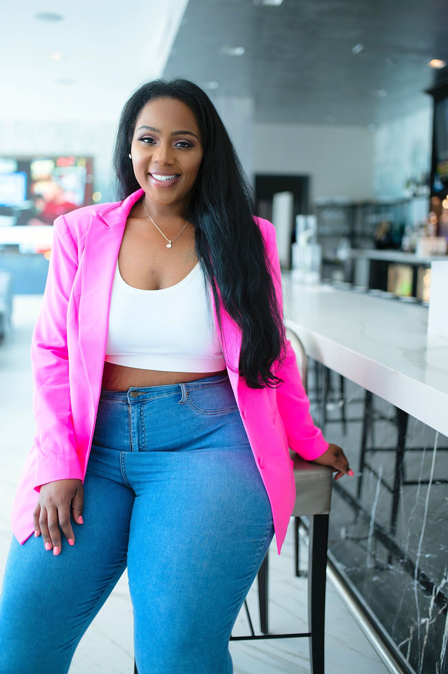Smiling woman in pink blazer and jeans leaning against bar counter in upscale venue.