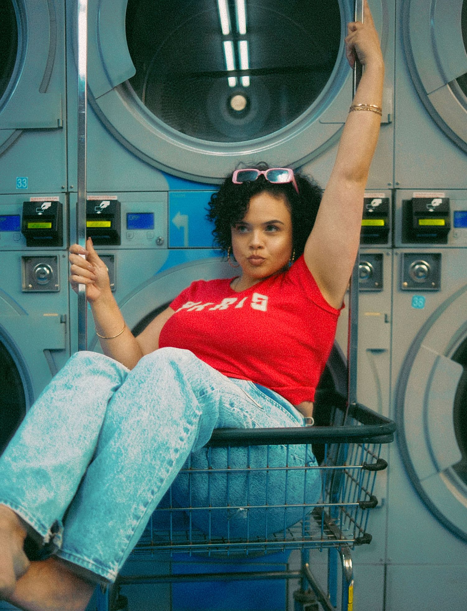 Young woman in red Paris tee and jeans sitting in laundry cart at laundromat.