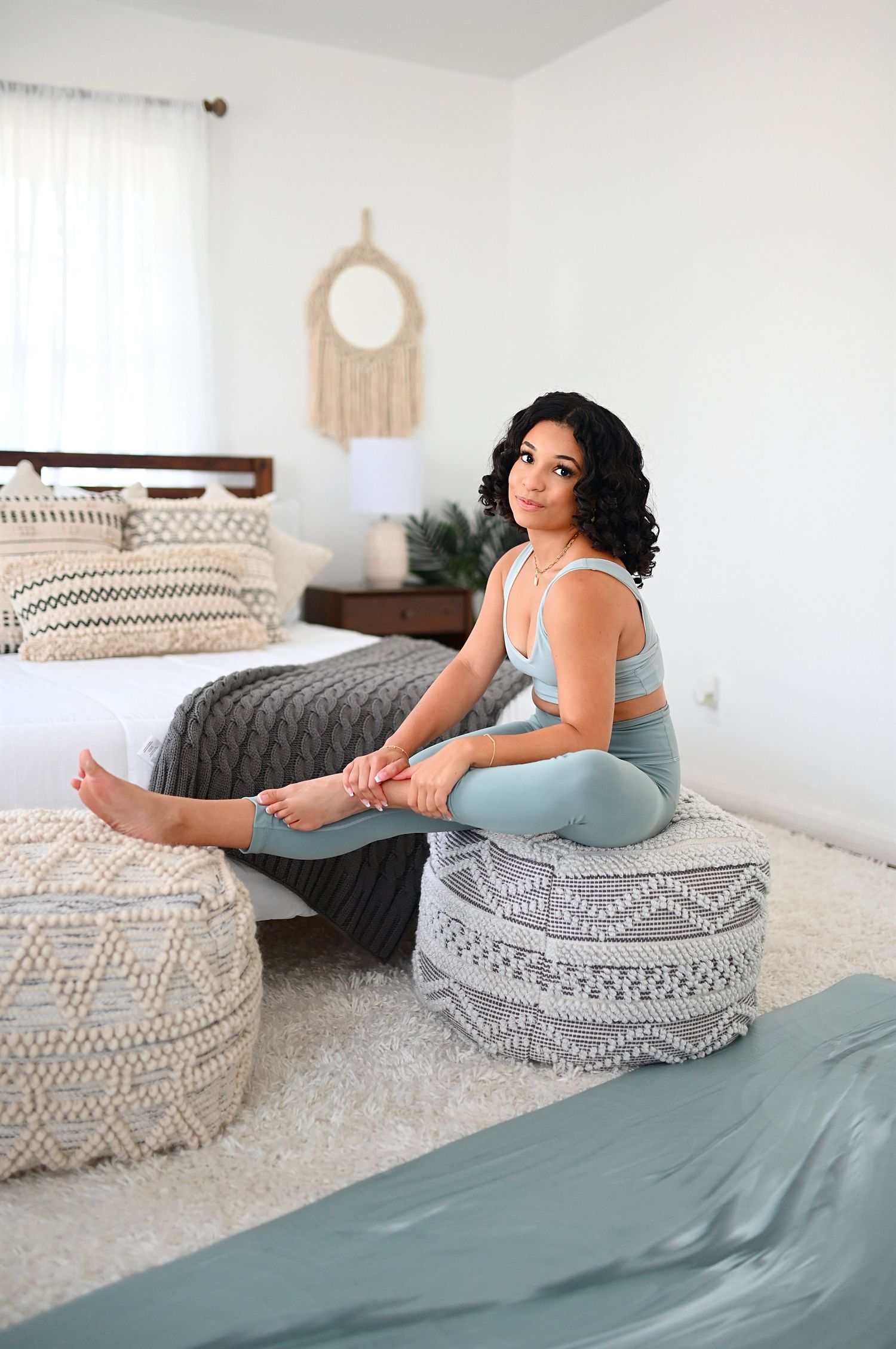 Woman in mint green sports bra and leggings sitting on woven pouf in boho bedroom.