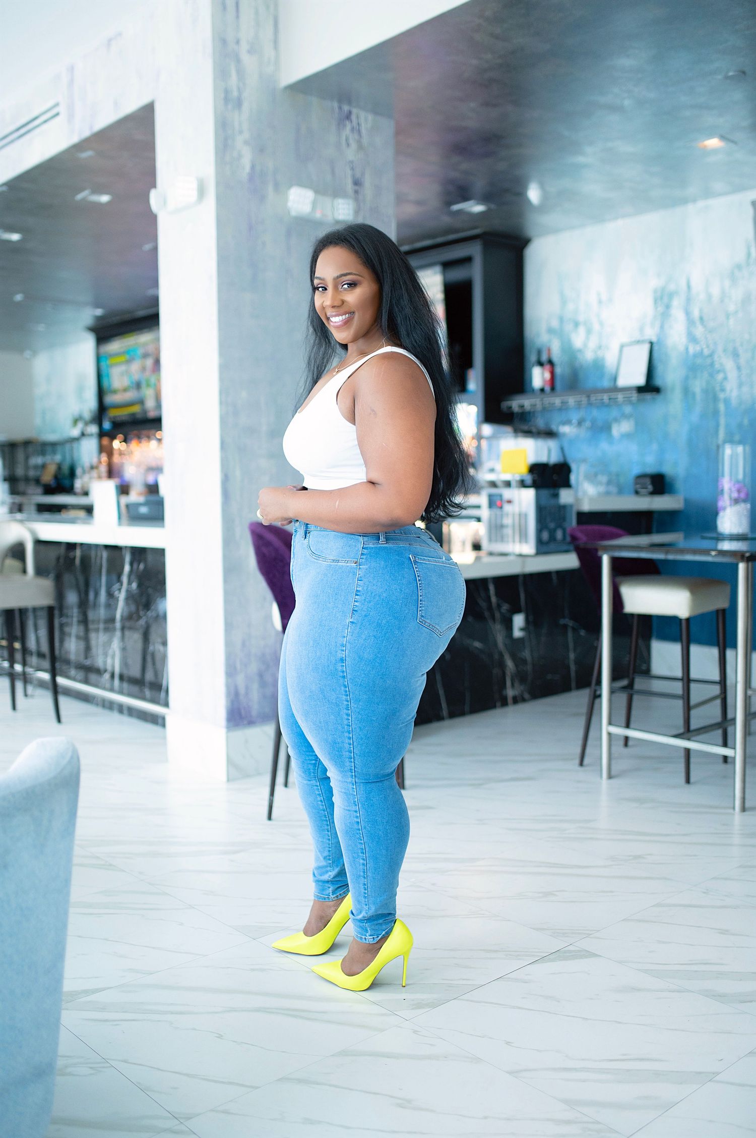 Woman in white crop top, jeans, and yellow heels posing in modern blue restaurant interior.