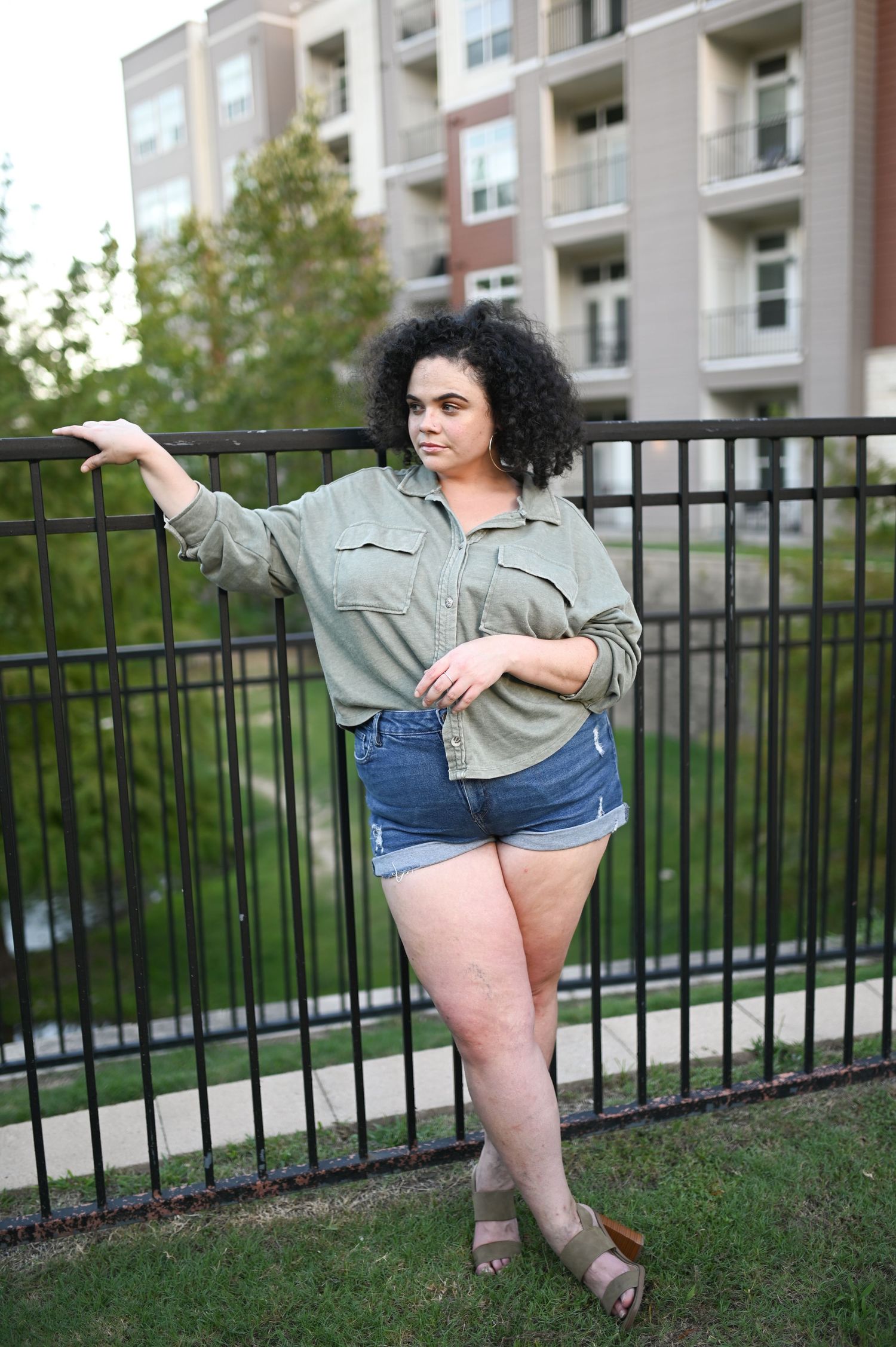 Curly-haired woman in olive shirt and denim shorts leans against black iron fence outdoors.
