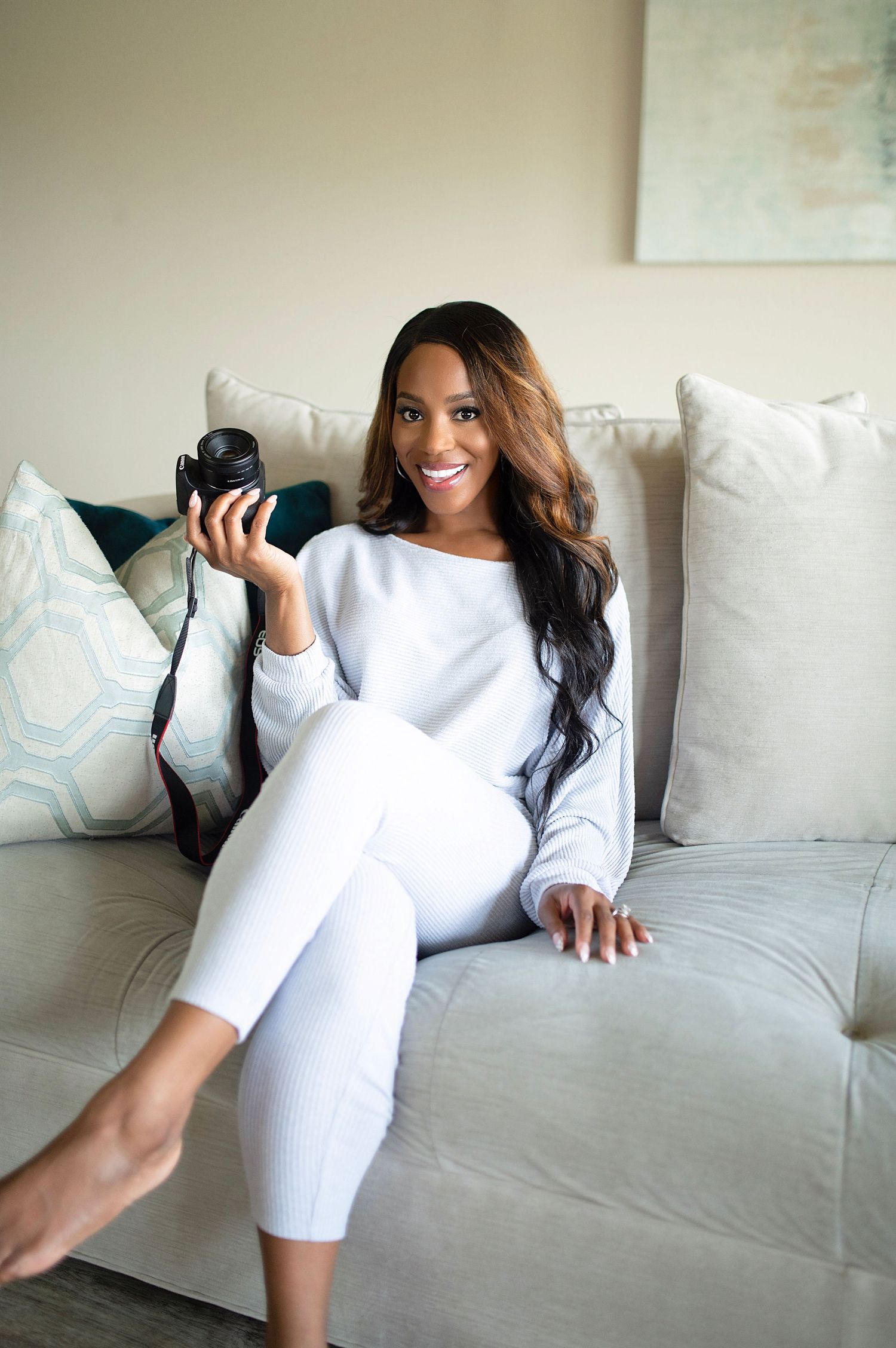 Smiling woman in white loungewear holds camera while sitting on neutral couch with decorative pillows.