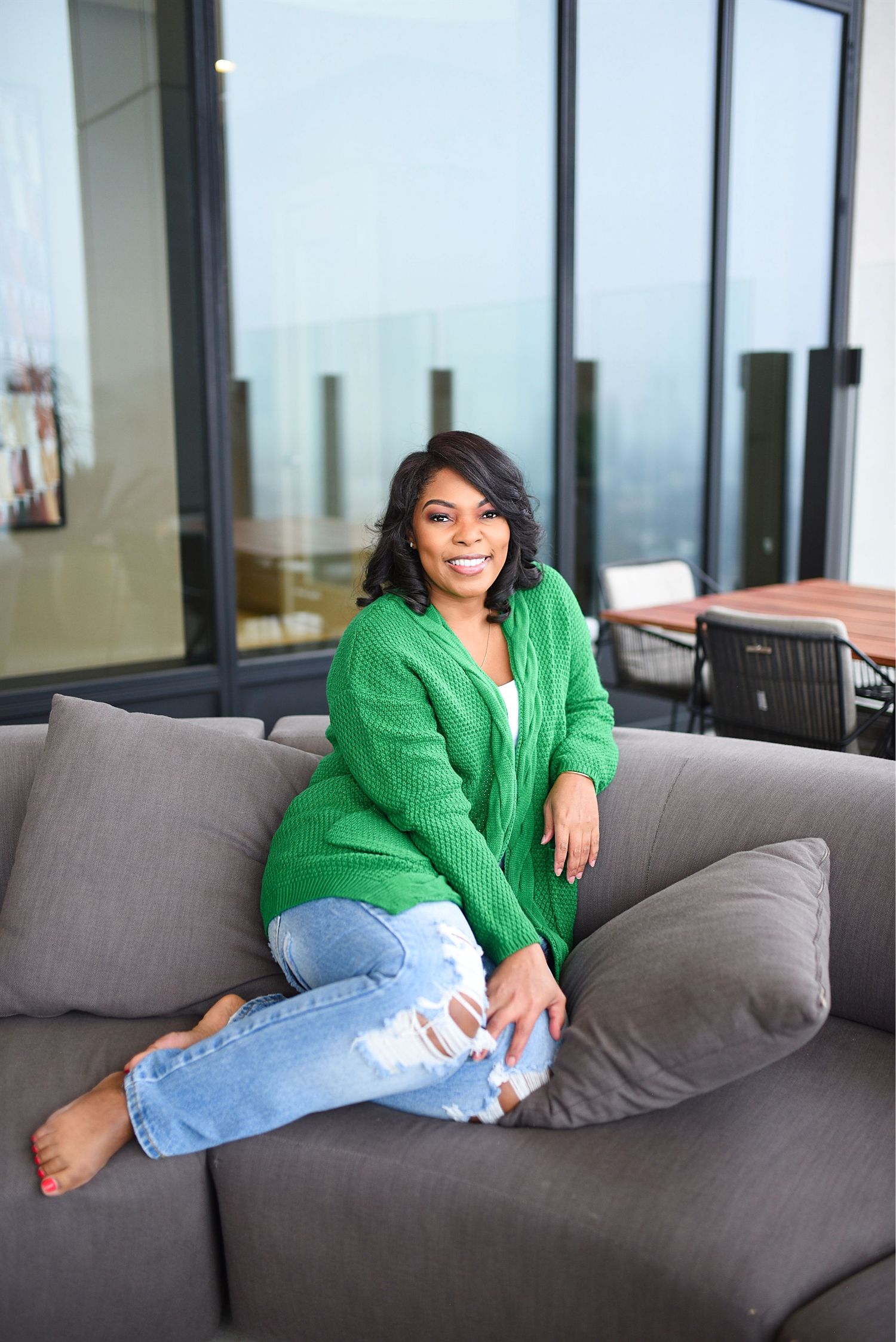 Smiling Black woman in green cardigan and ripped jeans sits relaxed on gray outdoor sofa.