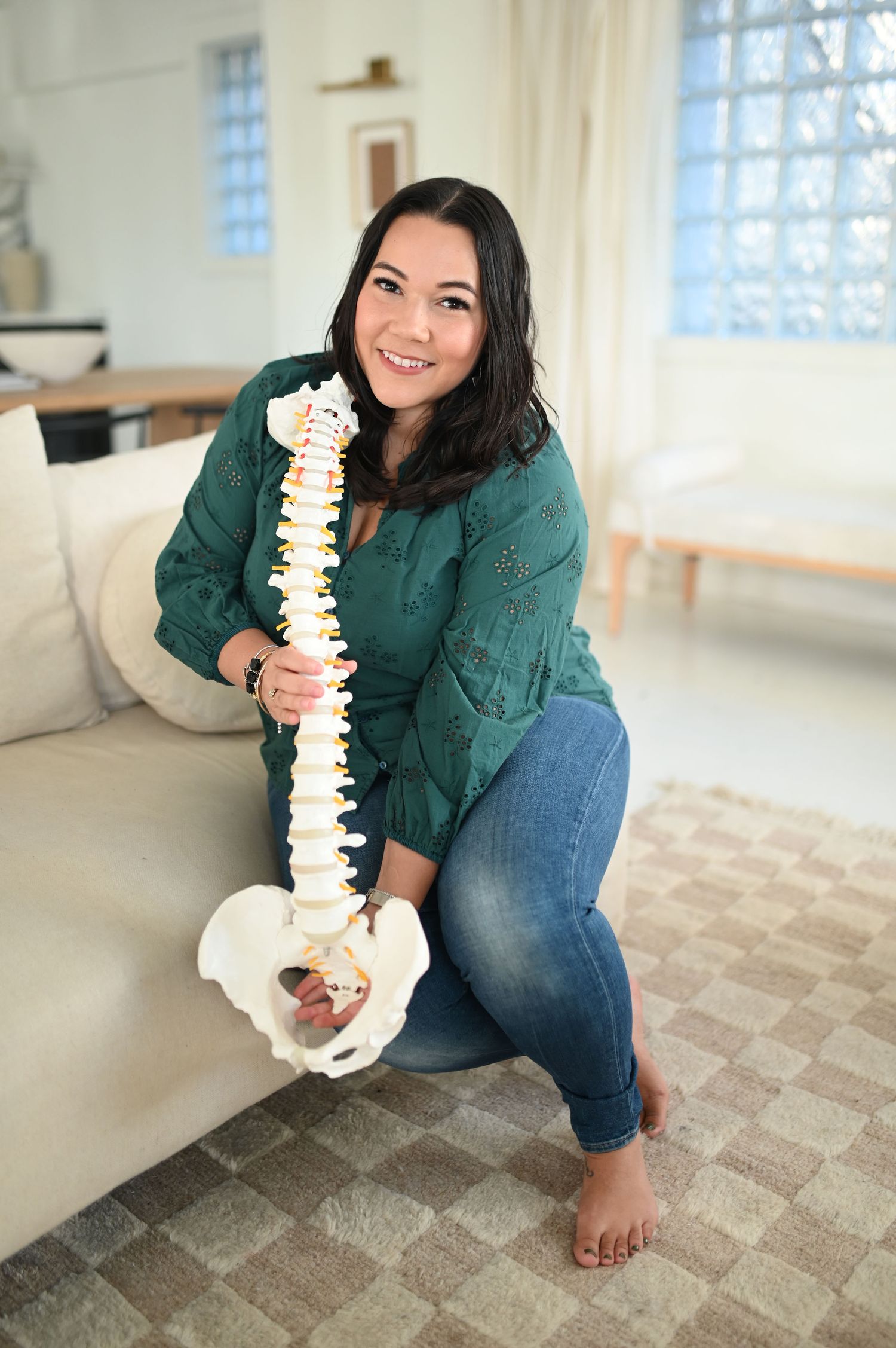 Smiling woman in green top and jeans posing with an anatomical spine model in a bright living room.