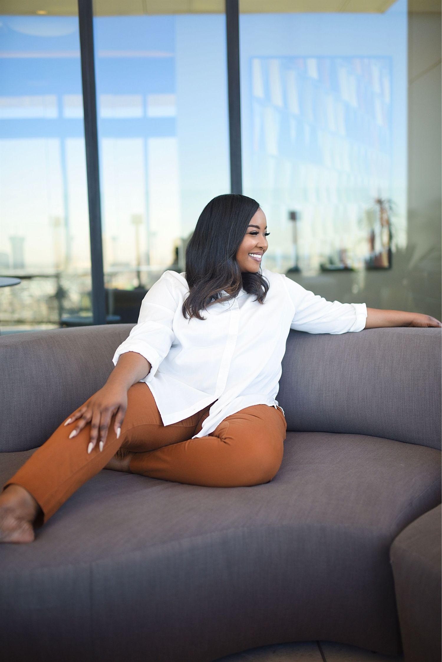 Smiling woman in white shirt and rust pants sits cross-legged on gray sofa near large windows.
