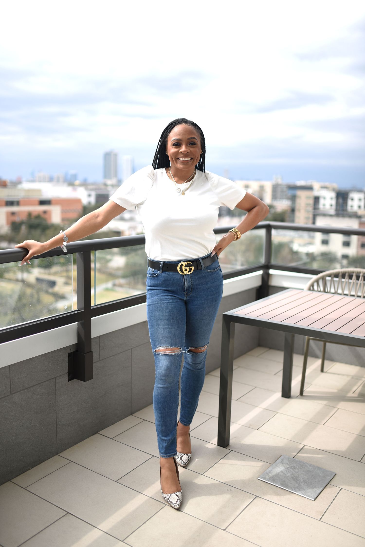 Smiling woman in white tee and ripped jeans with Gucci belt on rooftop balcony.