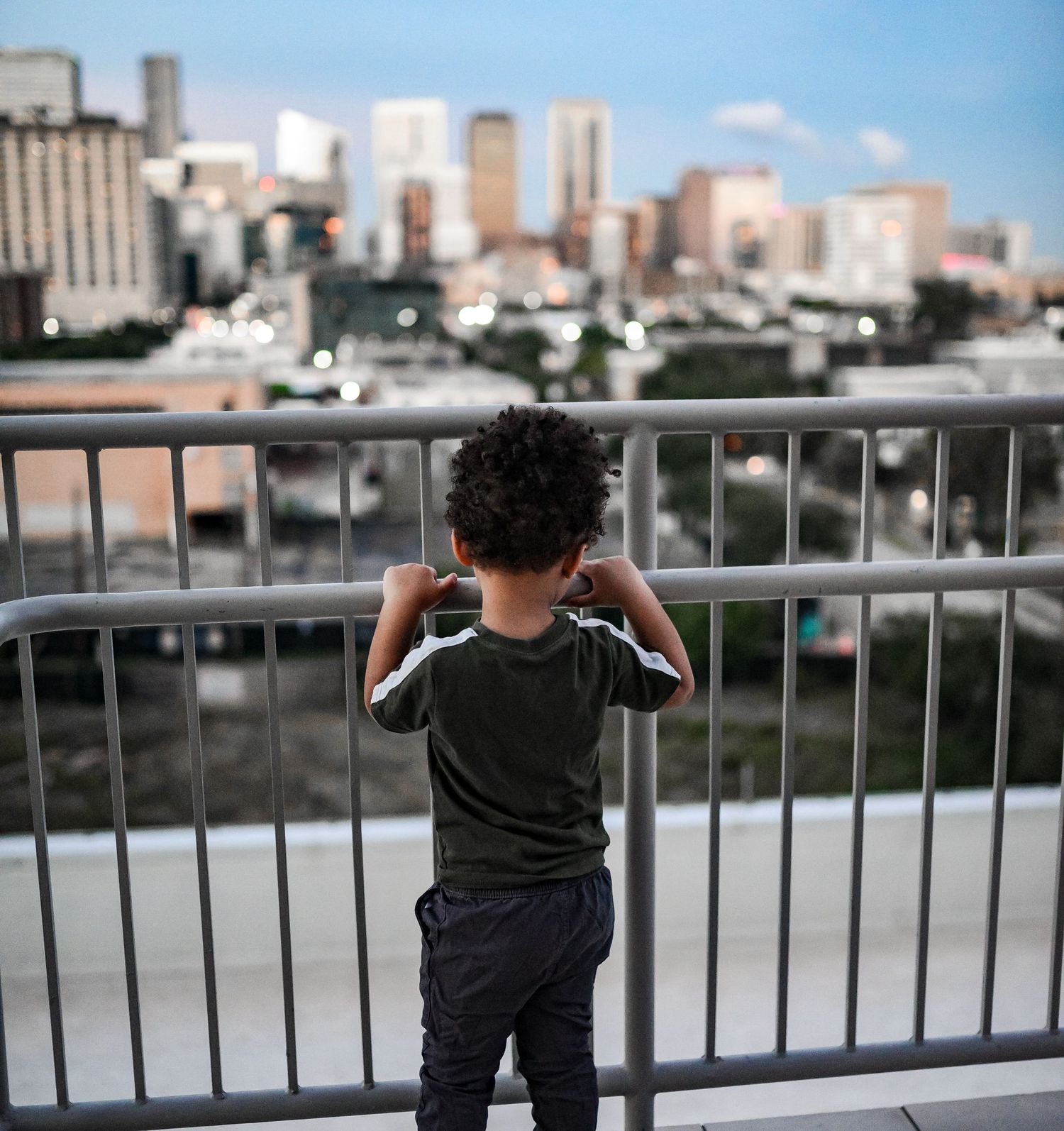 Young Black boy looking at city skyline through metal railing at dusk from overlook.