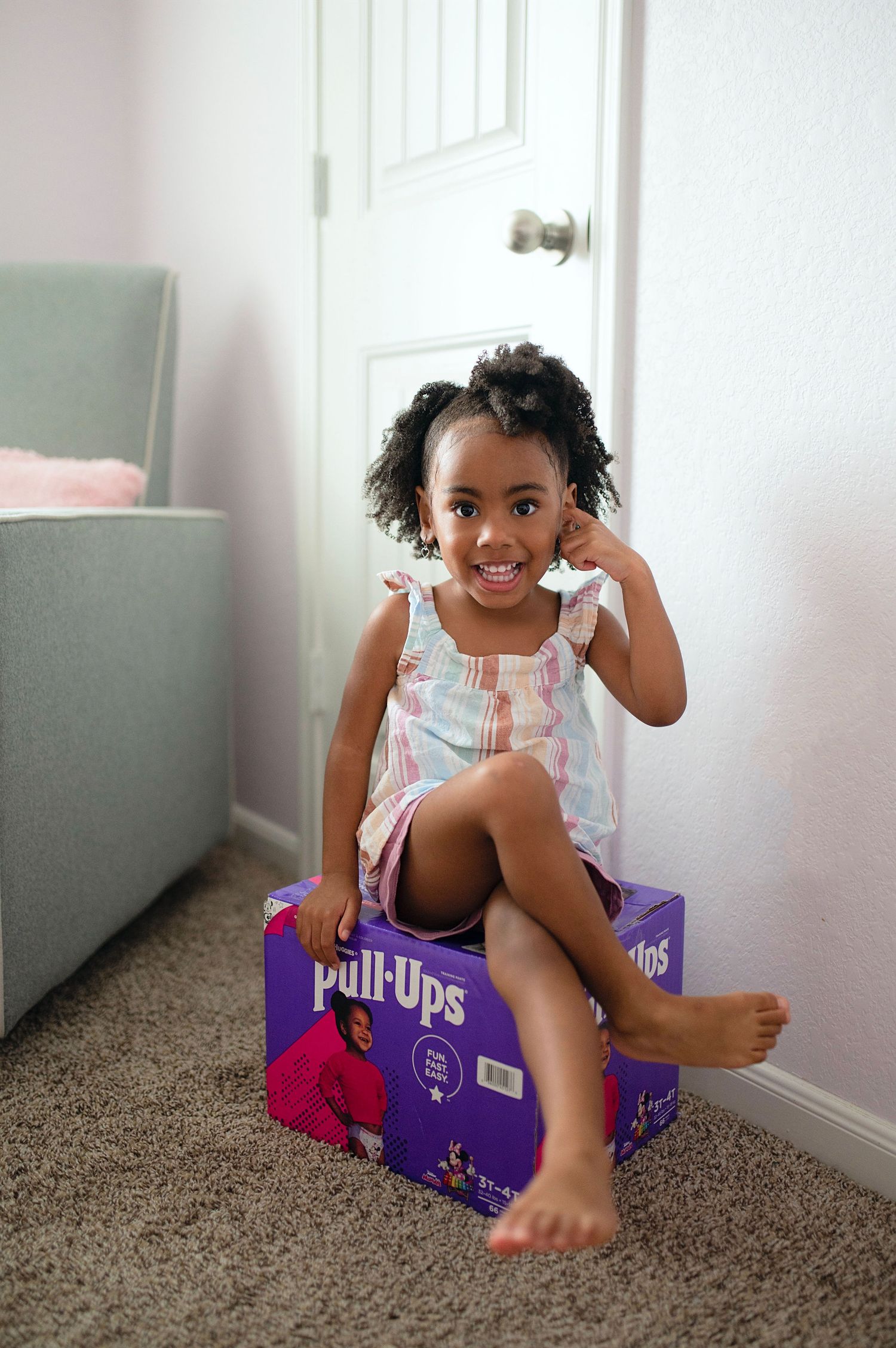 A cute young Black girl sits smiling on a Pull-Ups diaper box on carpet.