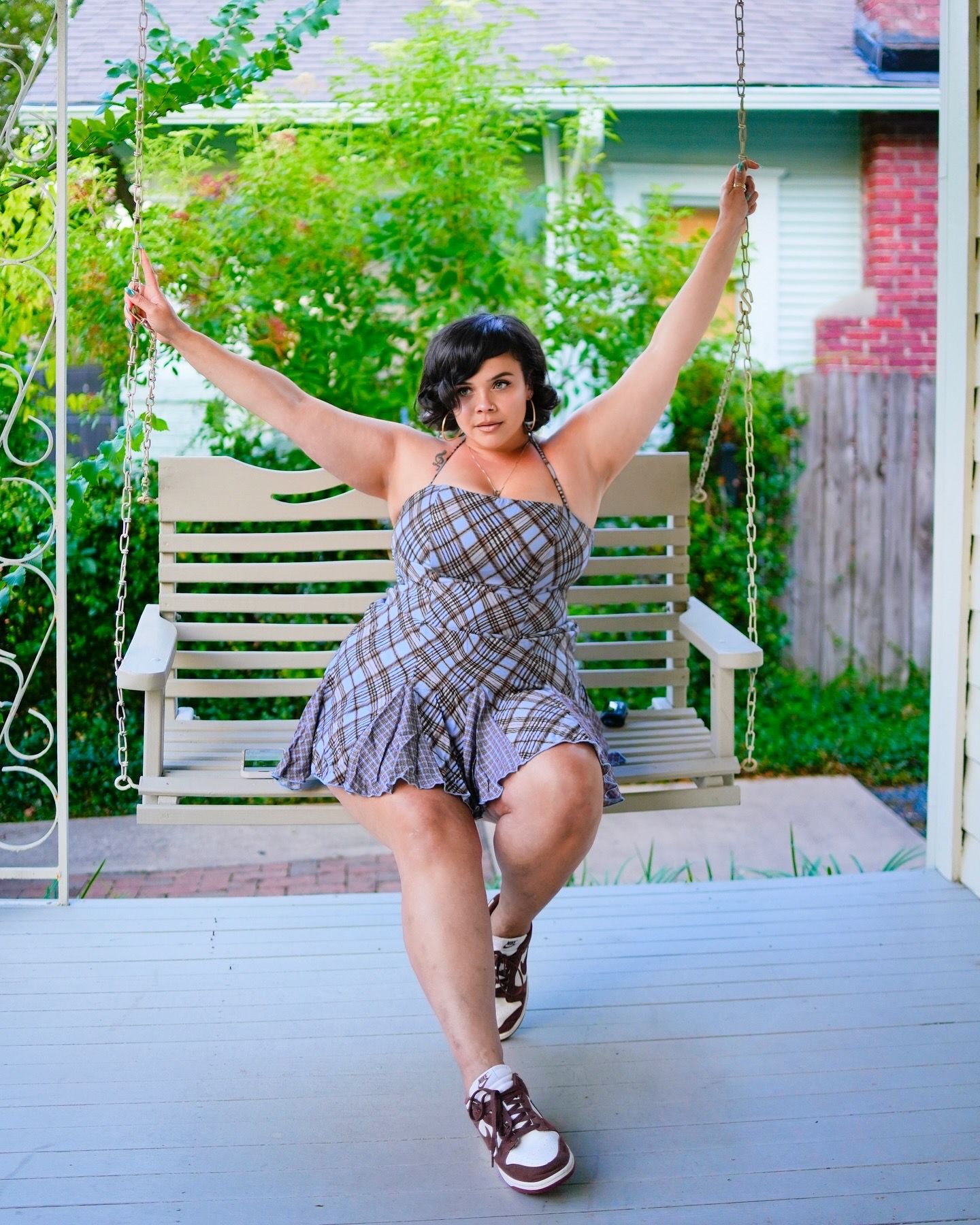 Woman in plaid mini dress poses playfully on porch swing with arms raised.