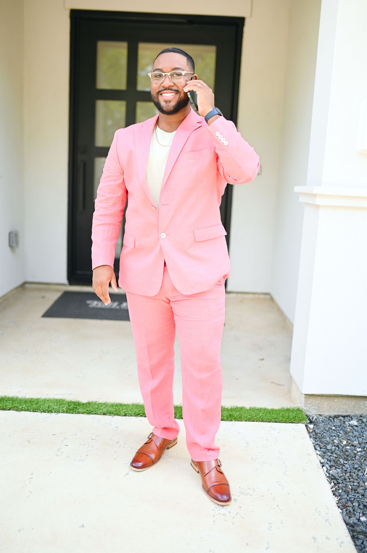 Smiling man in pink suit talking on phone while standing outside a white building.