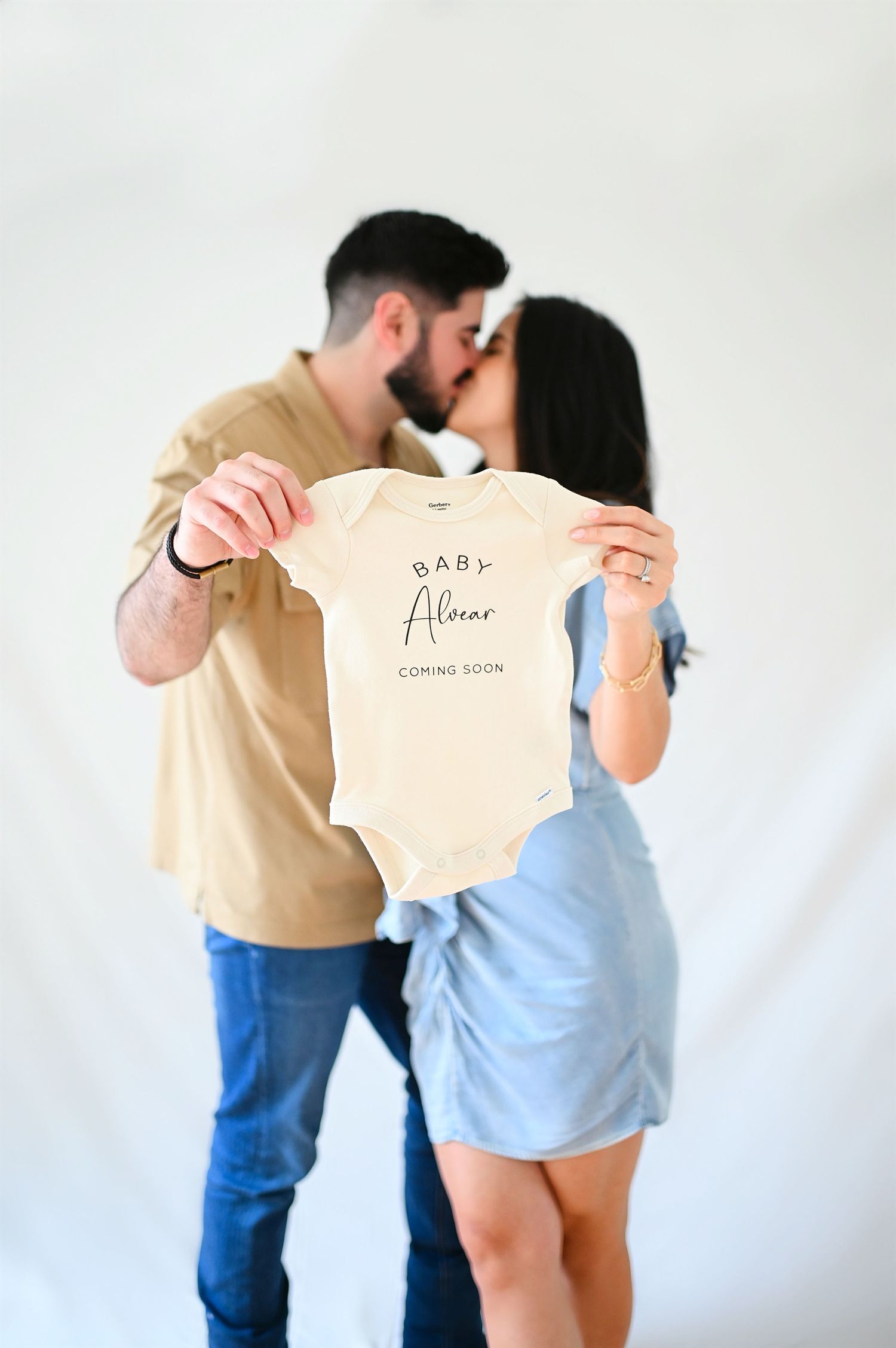 Couple kissing while holding Baby Alcazar coming soon onesie announcement.