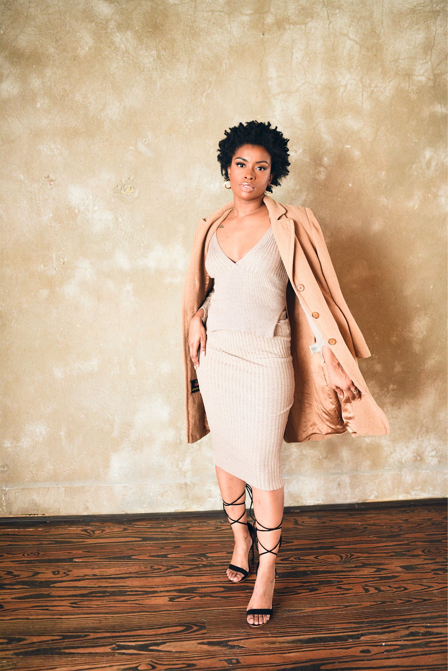 Woman in ribbed midi dress and camel coat walks confidently on wooden floor against textured backdrop.