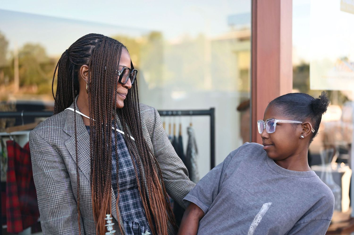 Two women with braids and glasses share a conversation outside a clothing store.