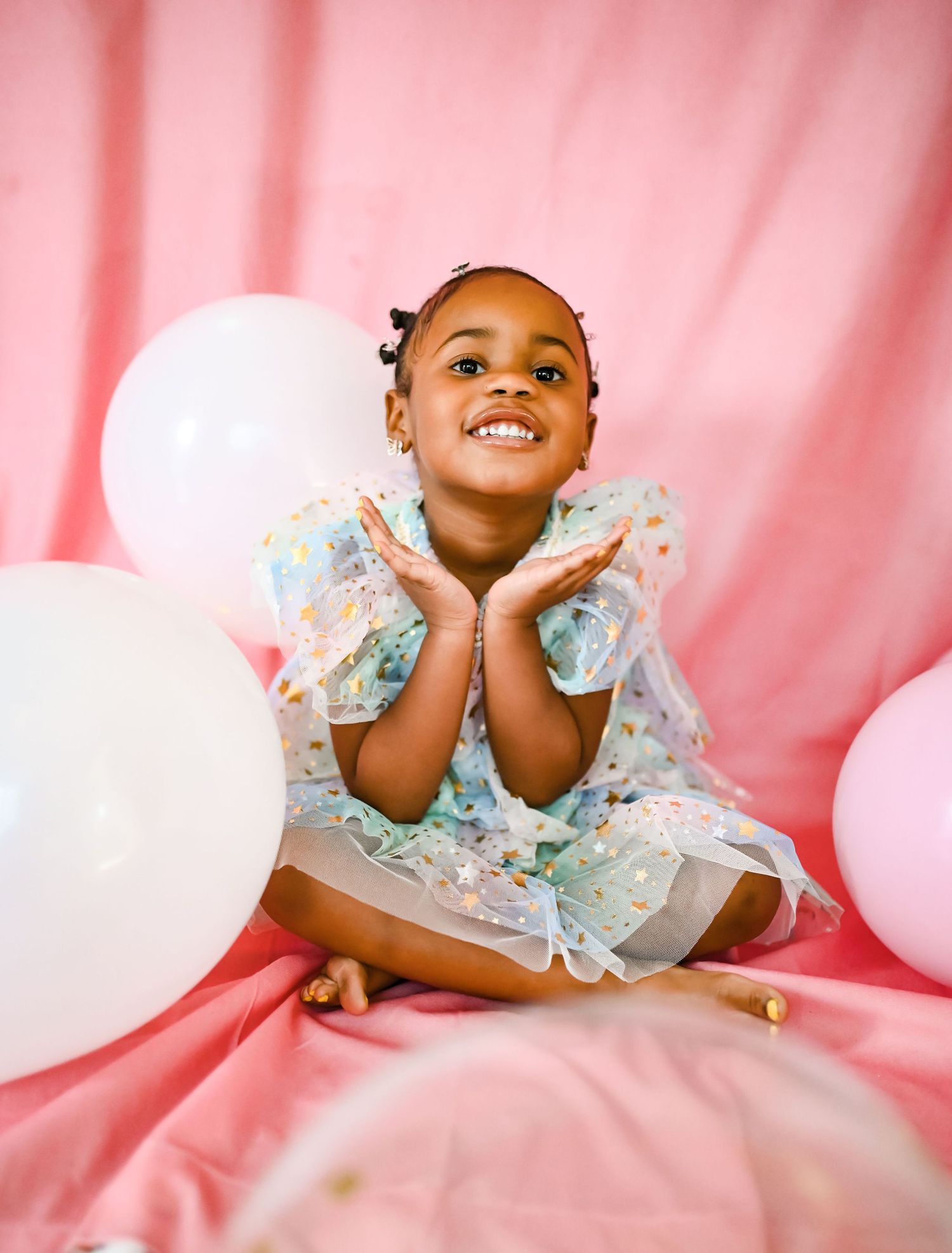 A joyful Black toddler girl in a sparkly dress sits among pink and white balloons.