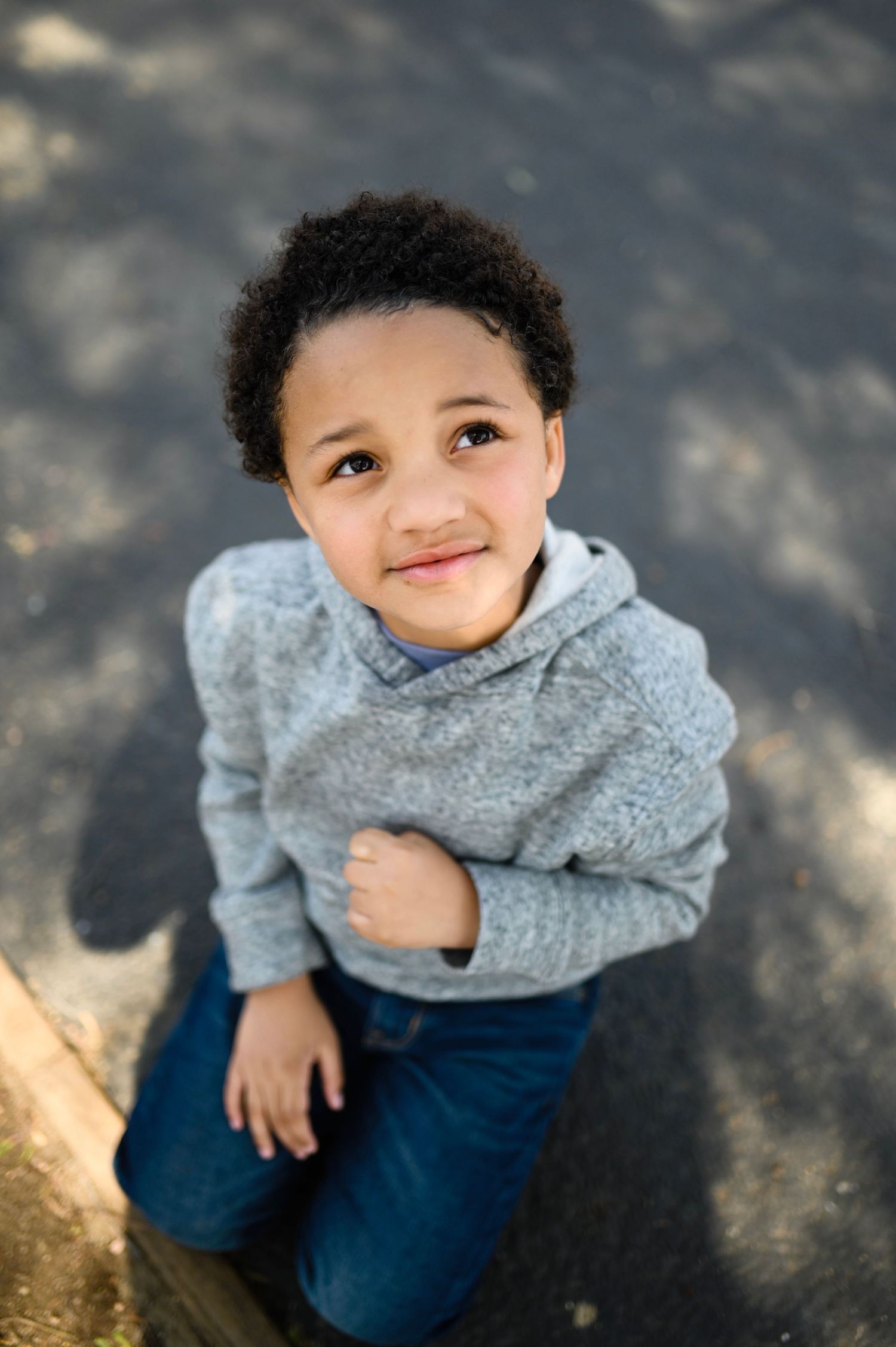 Cute curly-haired boy in gray hoodie and jeans sits outdoors looking up at camera.