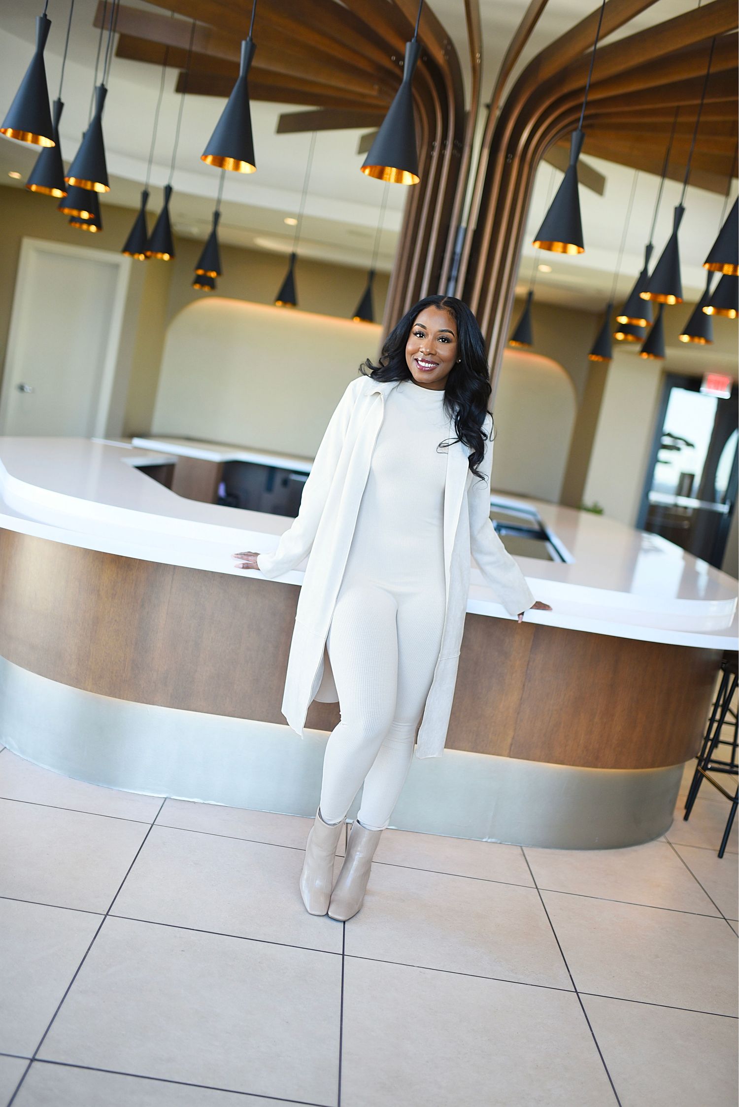 Woman in all-white outfit smiling while leaning on a curved reception desk with pendant lights.