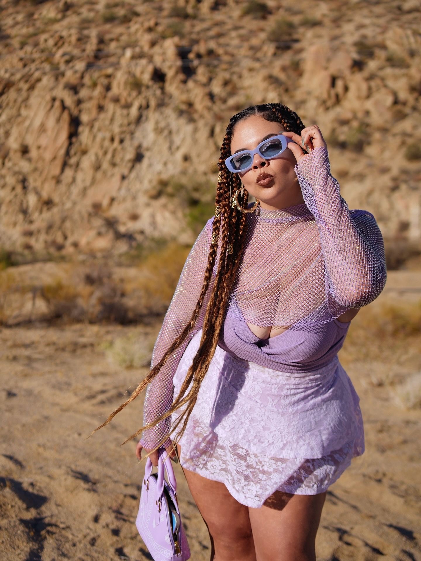 Woman with long braids in purple mesh outfit adjusts sunglasses in desert setting.