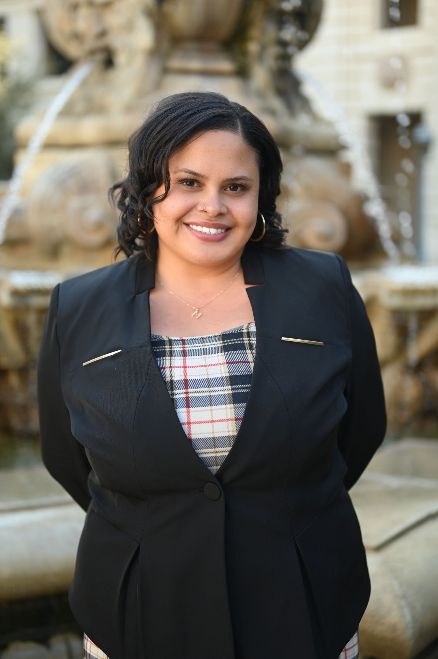 Professional woman in black blazer and plaid dress smiling in front of decorative fountain outdoors.