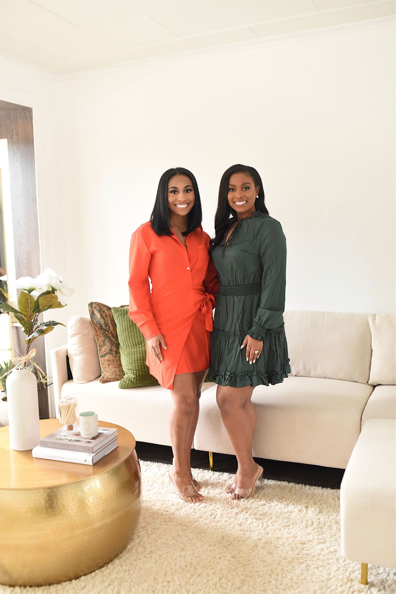 Two women in orange and green mini dresses standing together in bright modern living room.