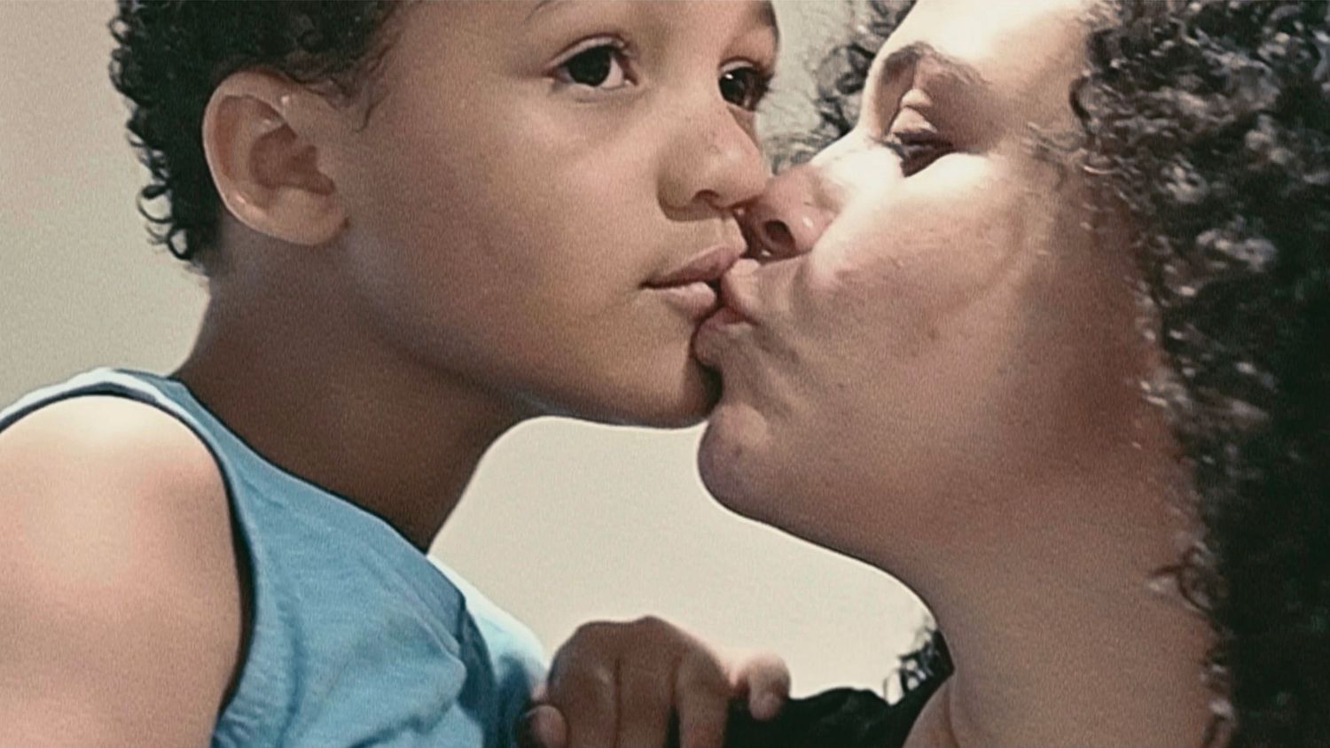 A mother tenderly kisses her young curly-haired son on the cheek in a warm, intimate moment.