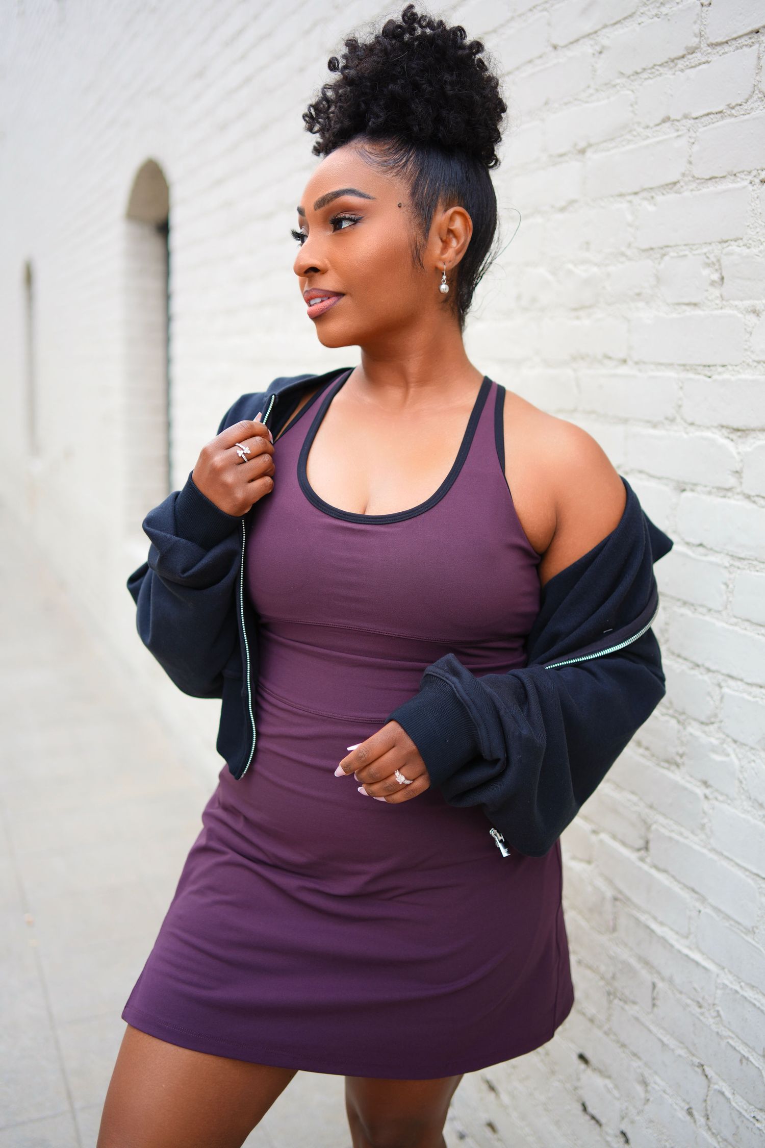 Black woman in purple athletic dress and navy zip hoodie posing near white brick wall.
