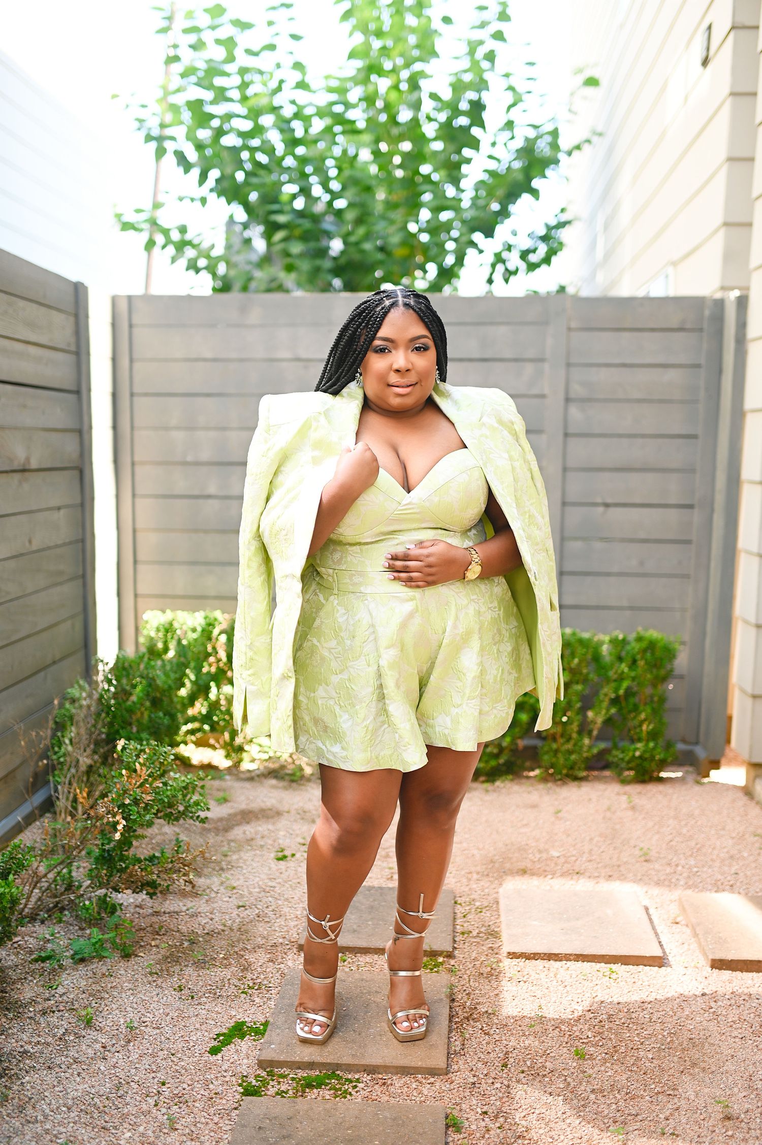 Plus-size woman posing in a lime green floral romper and matching blazer with strappy heels outdoors.