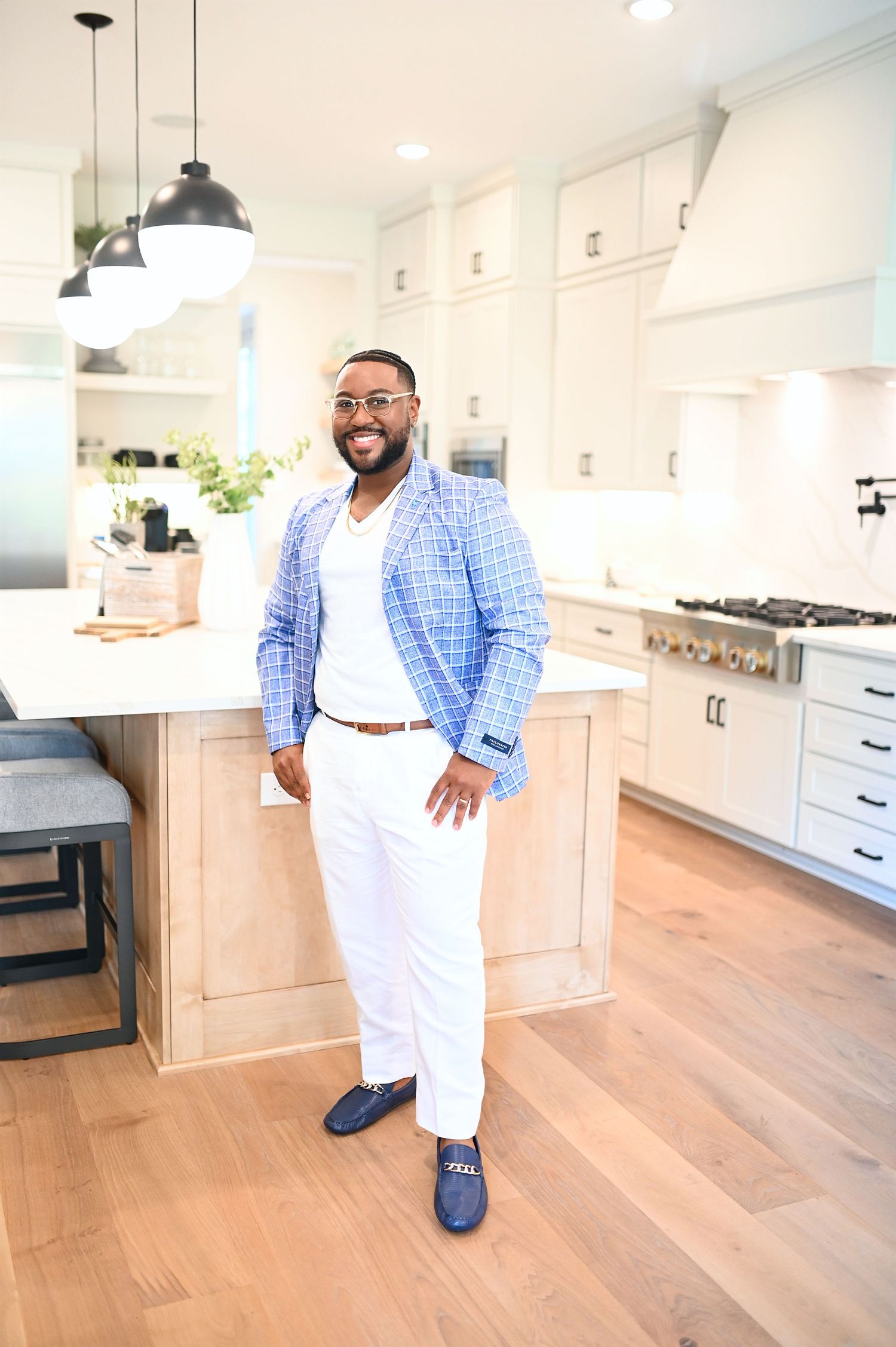 Stylish man in blue plaid blazer and white pants posing in a modern white kitchen.