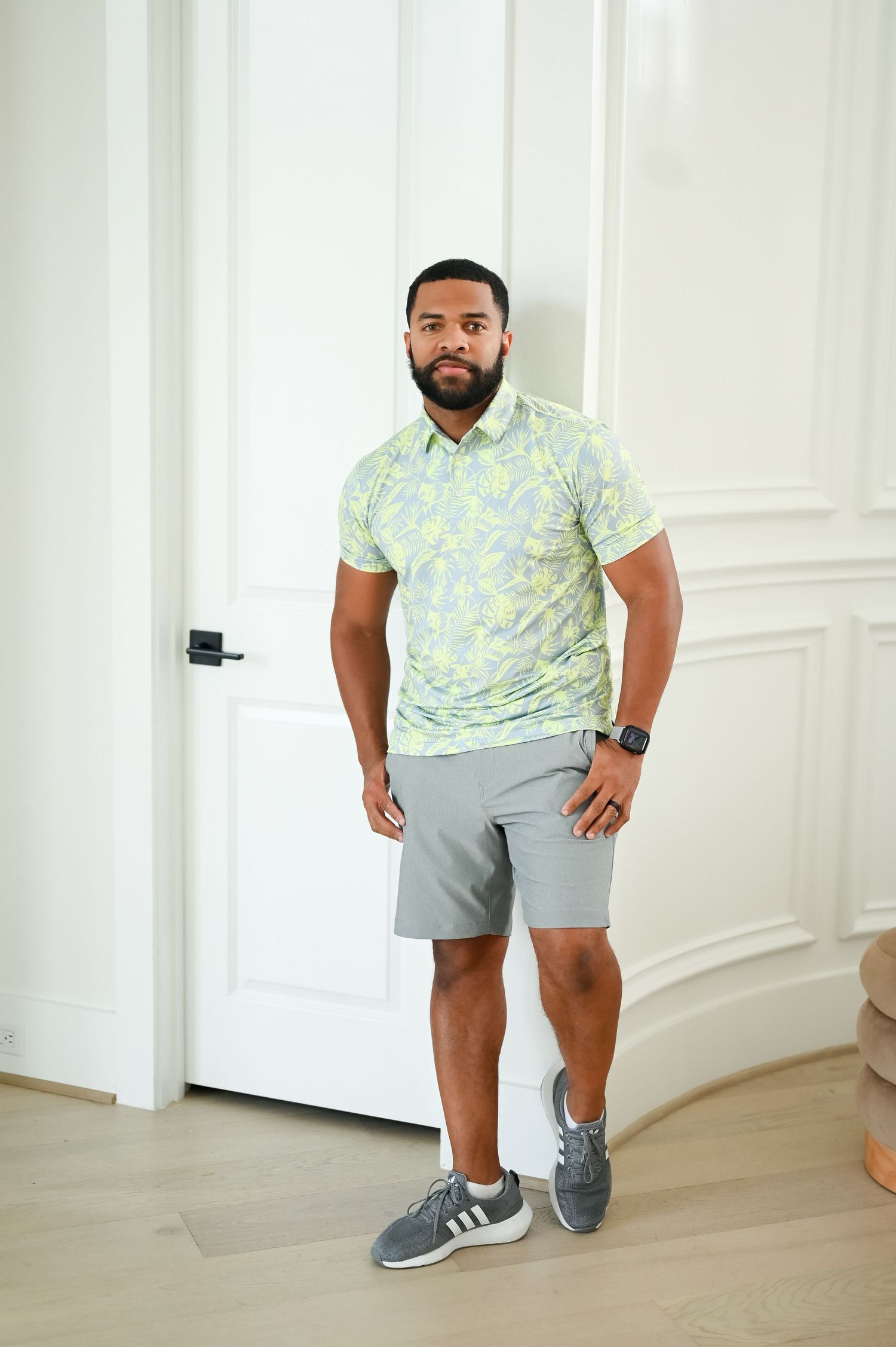 Athletic man in yellow patterned polo shirt and gray shorts wearing Adidas sneakers posing indoors.