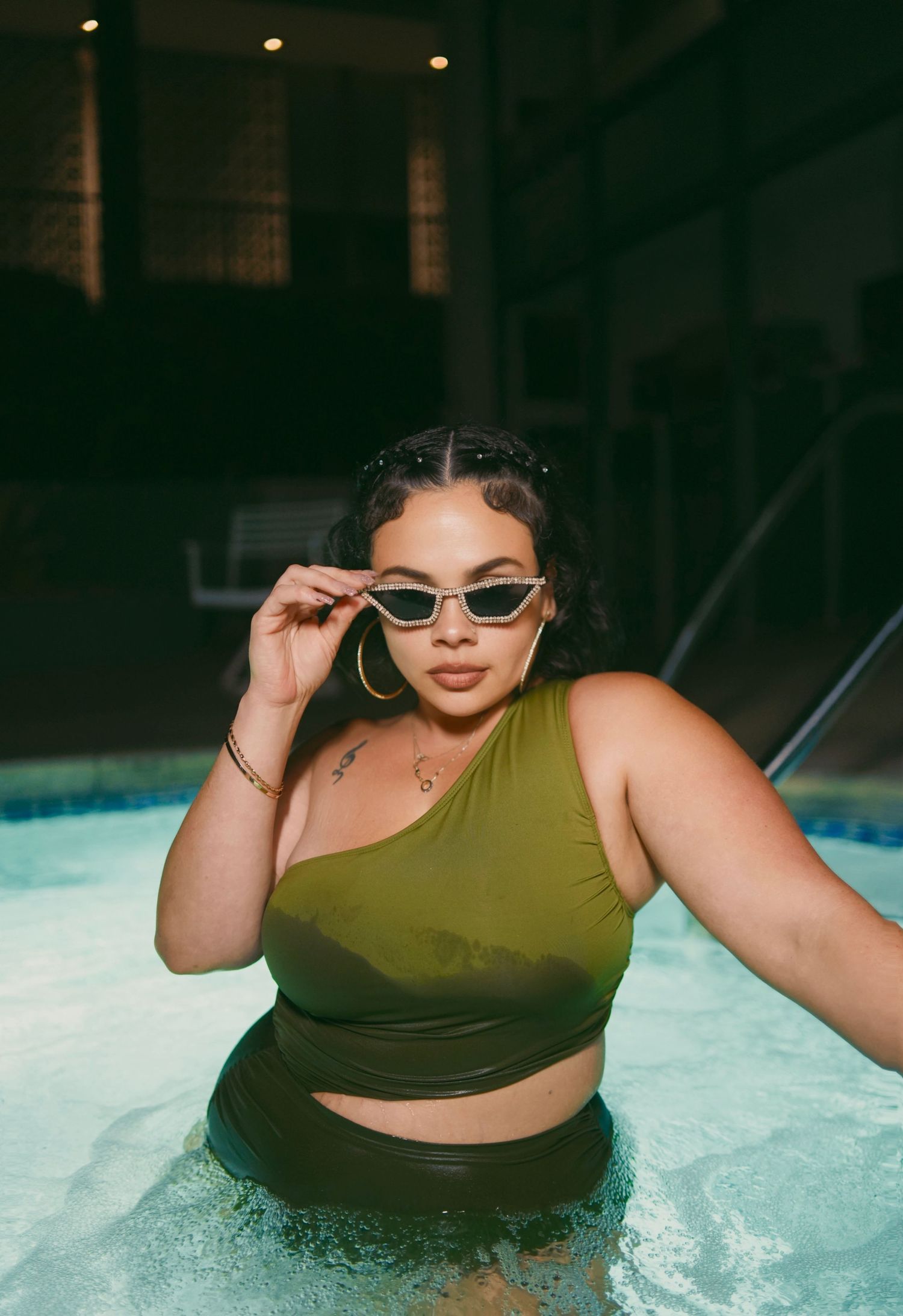 Woman in olive green one-shoulder bikini top poses in pool wearing small cat-eye sunglasses and gold hoop earrings.