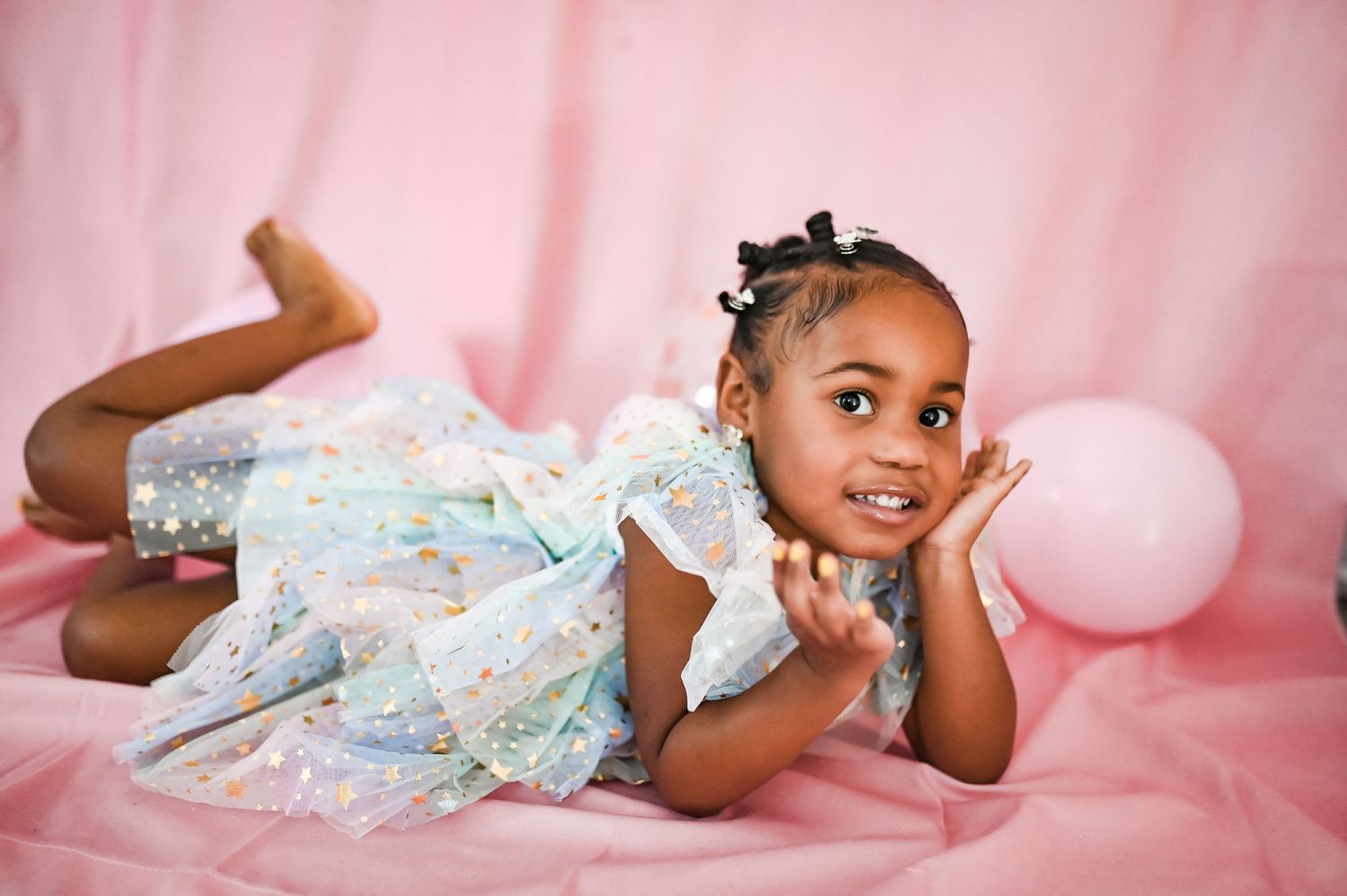 An adorable Black toddler girl in a sparkly dress poses on a pink backdrop.
