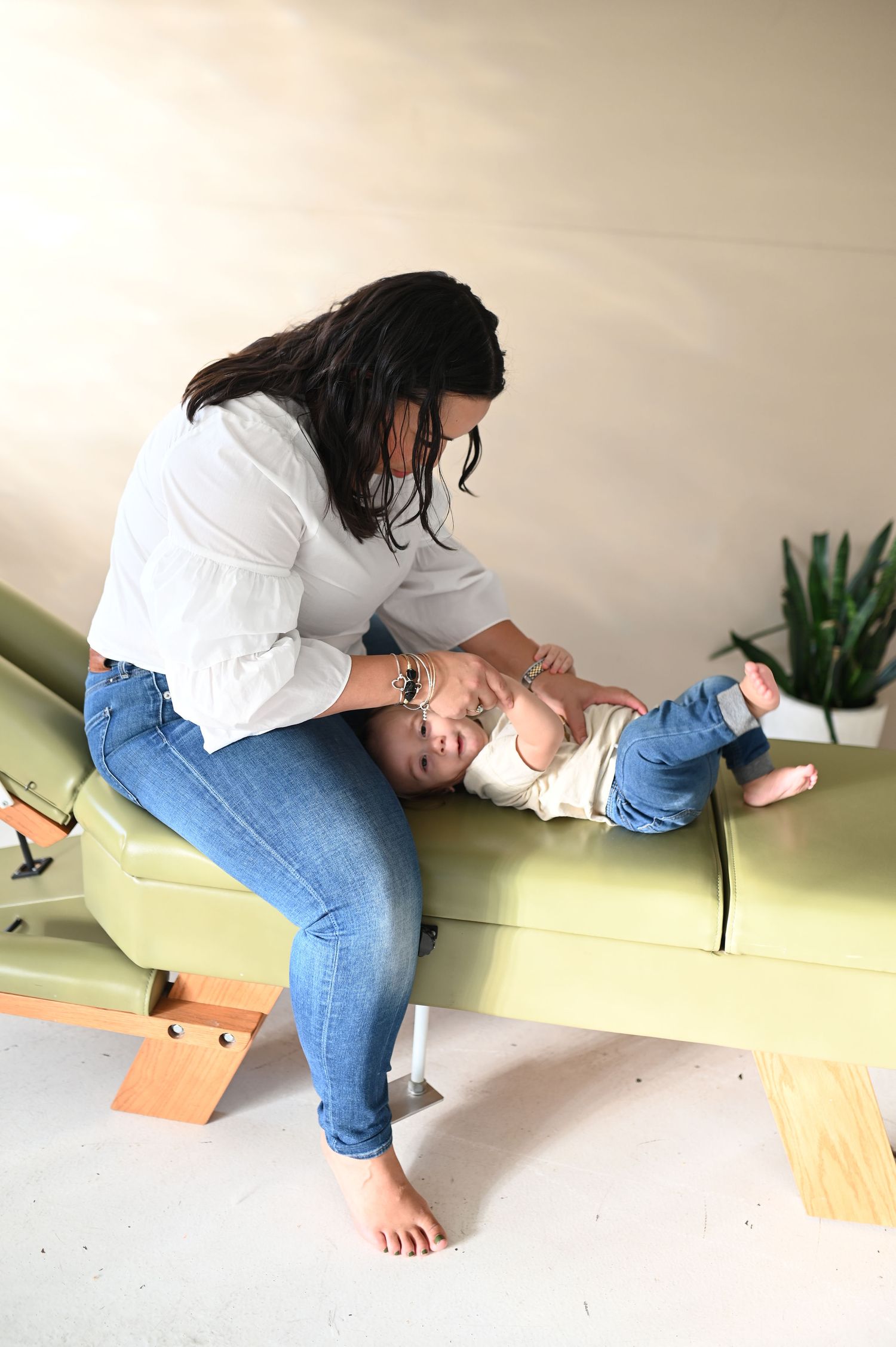 Woman in white shirt and jeans plays with baby on green chiropractic table indoors.