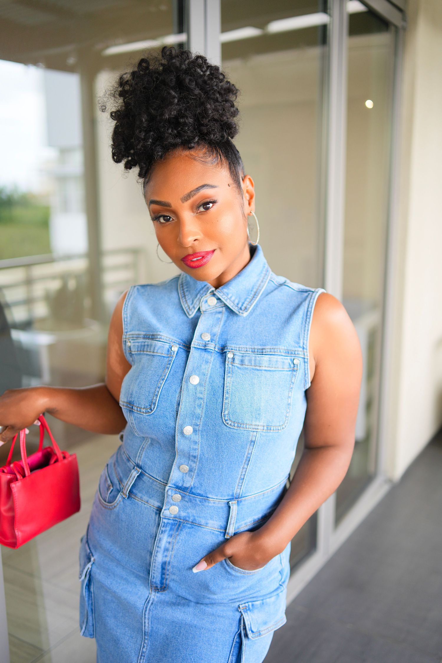 Black woman in sleeveless denim dress holding red handbag posing outside glass building.