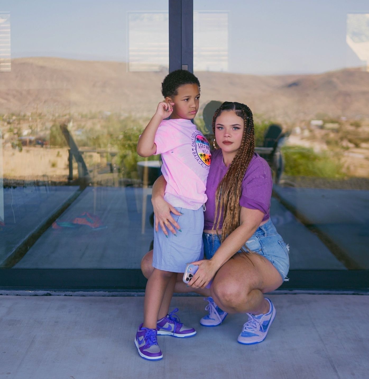 Young boy and woman in purple top and denim shorts posing together in front of large glass windows outdoors.