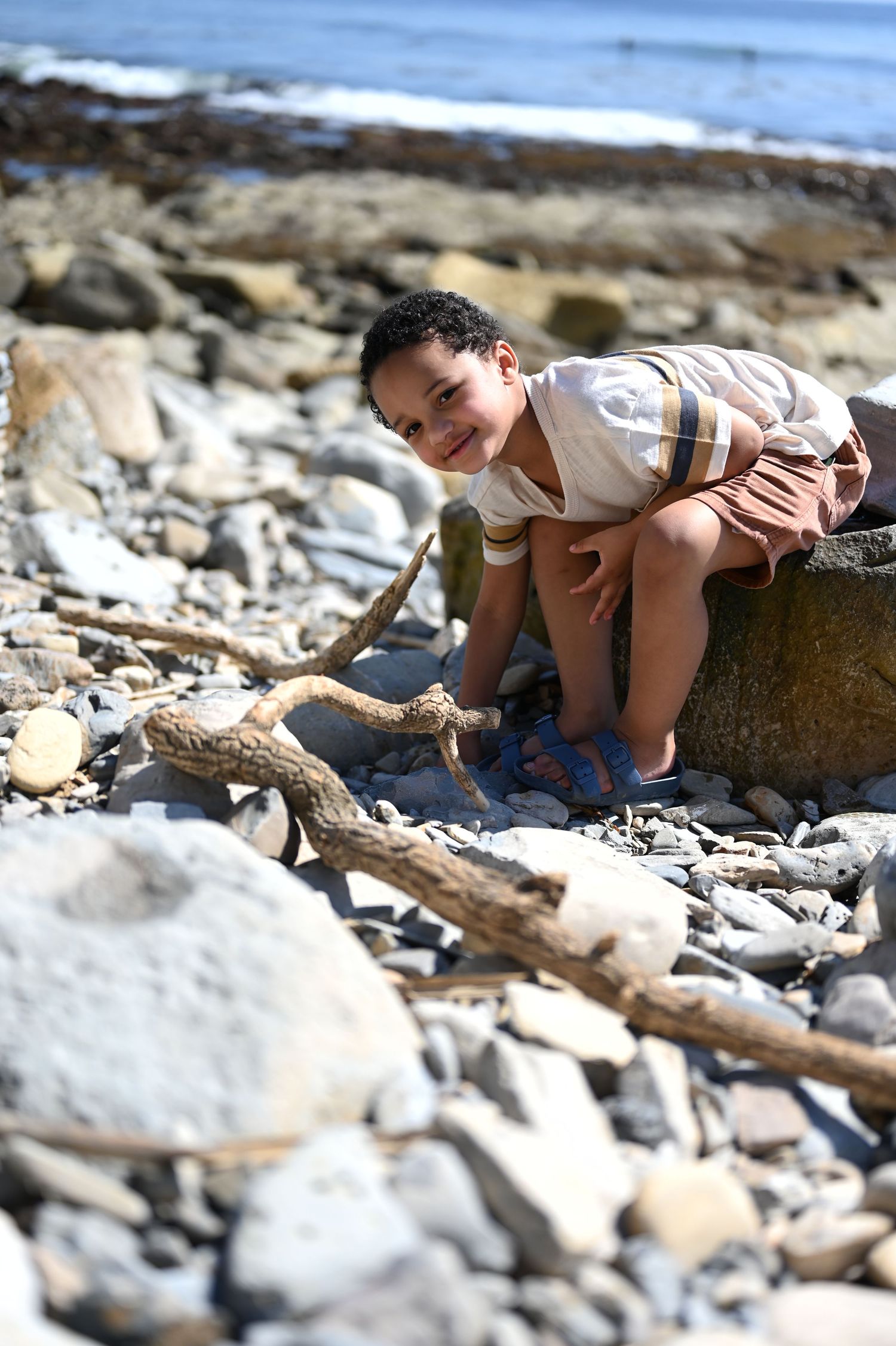 Young Black boy playing on rocky beach with driftwood, smiling near ocean on sunny day.