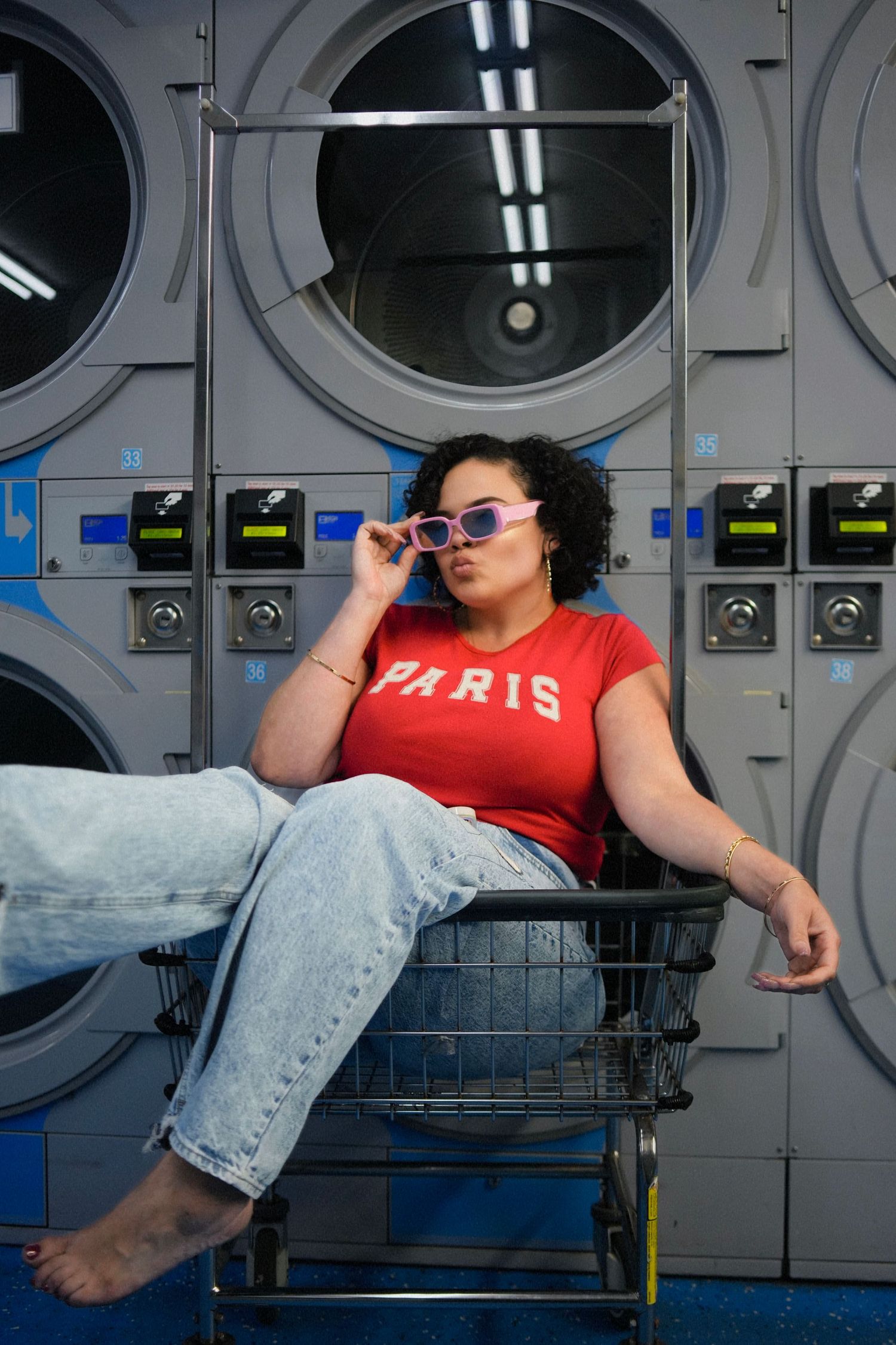 Woman in red Paris shirt and jeans posing in laundry basket at laundromat.