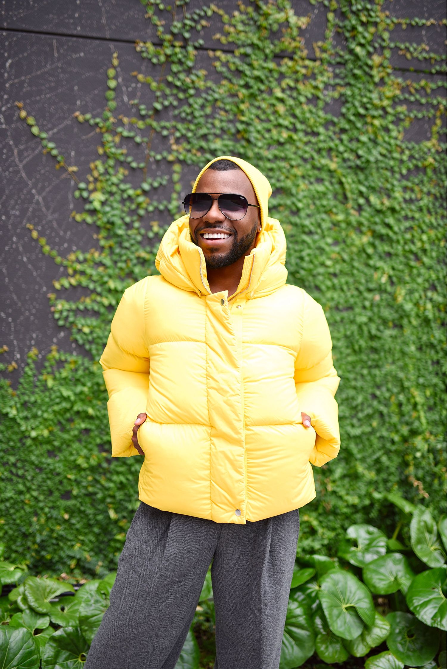 Man in bright yellow puffer jacket and sunglasses smiling in front of ivy-covered wall.