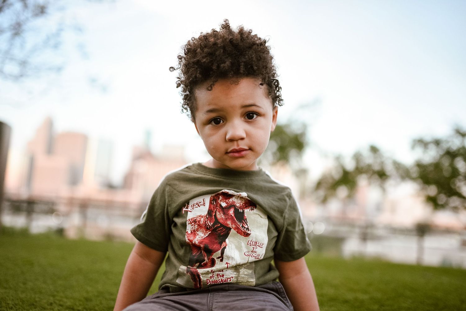 Young curly-haired boy in olive graphic tee sits outdoors with city skyline behind him.