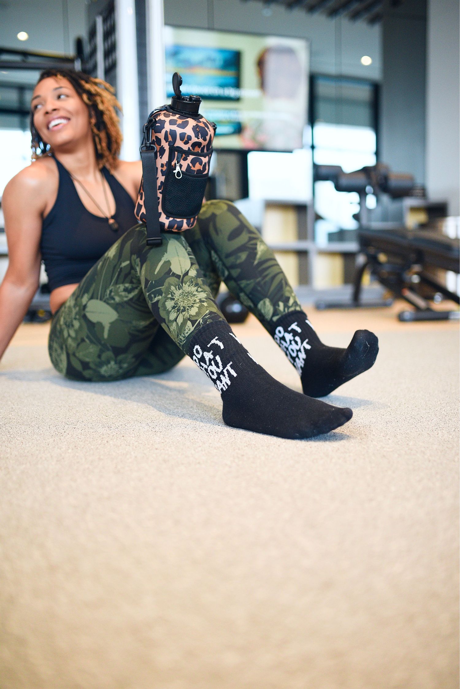 Smiling woman in camo leggings and black top sitting on gym floor with leopard print bag on her leg.